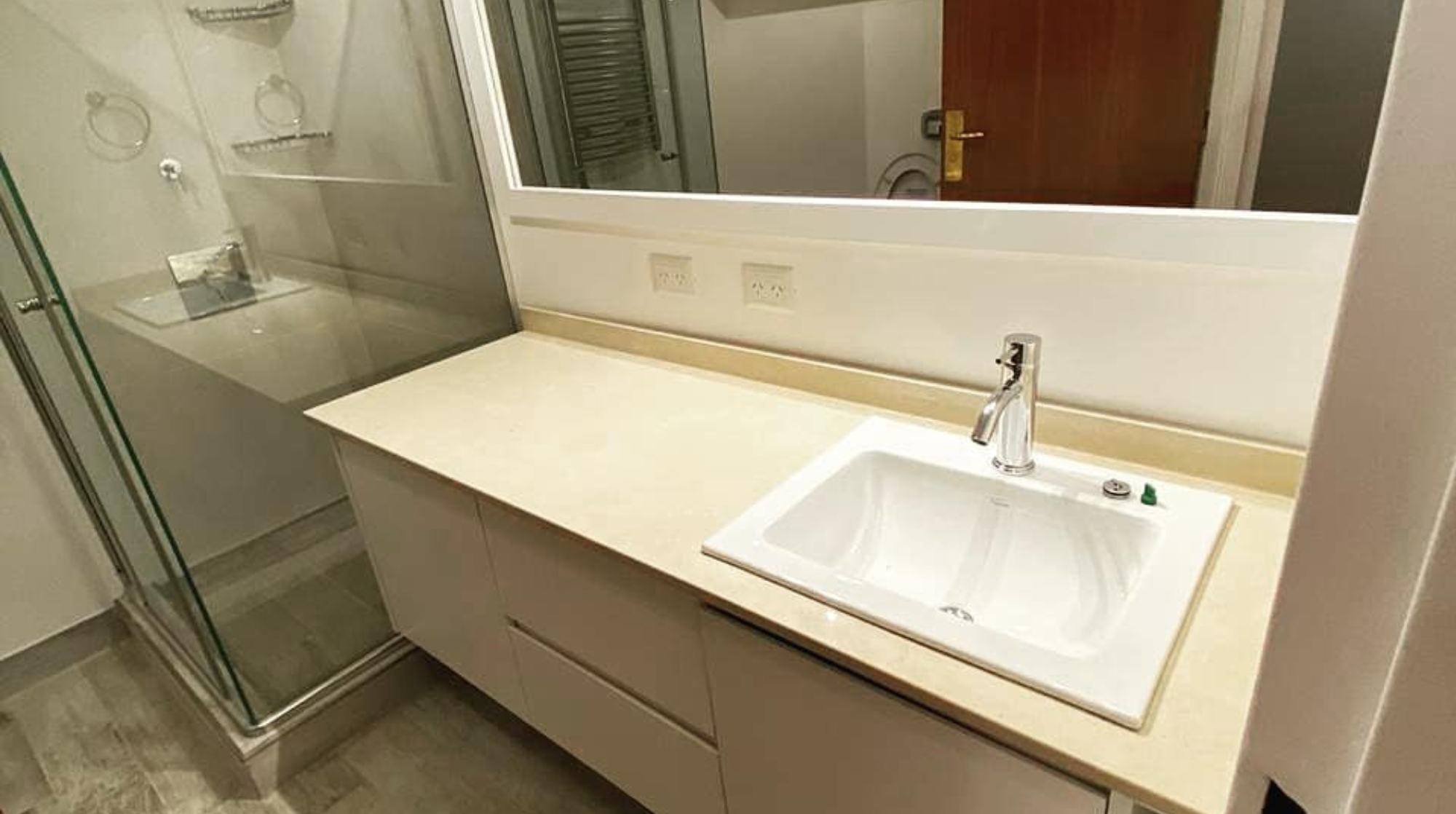 Lavabo con fregadero en un baño, con espejo grande, dos enchufes y una ducha con puerta de cristal en el fondo.