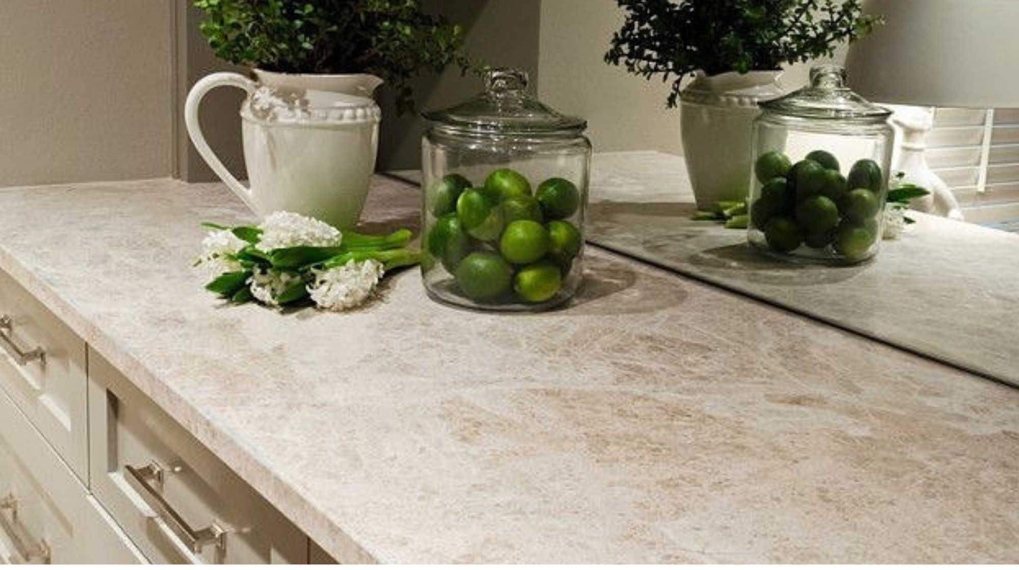Una mesa de cocina con botellas de vidrio con limones, un florero con flores blancas y jarrones con plantas verdes.