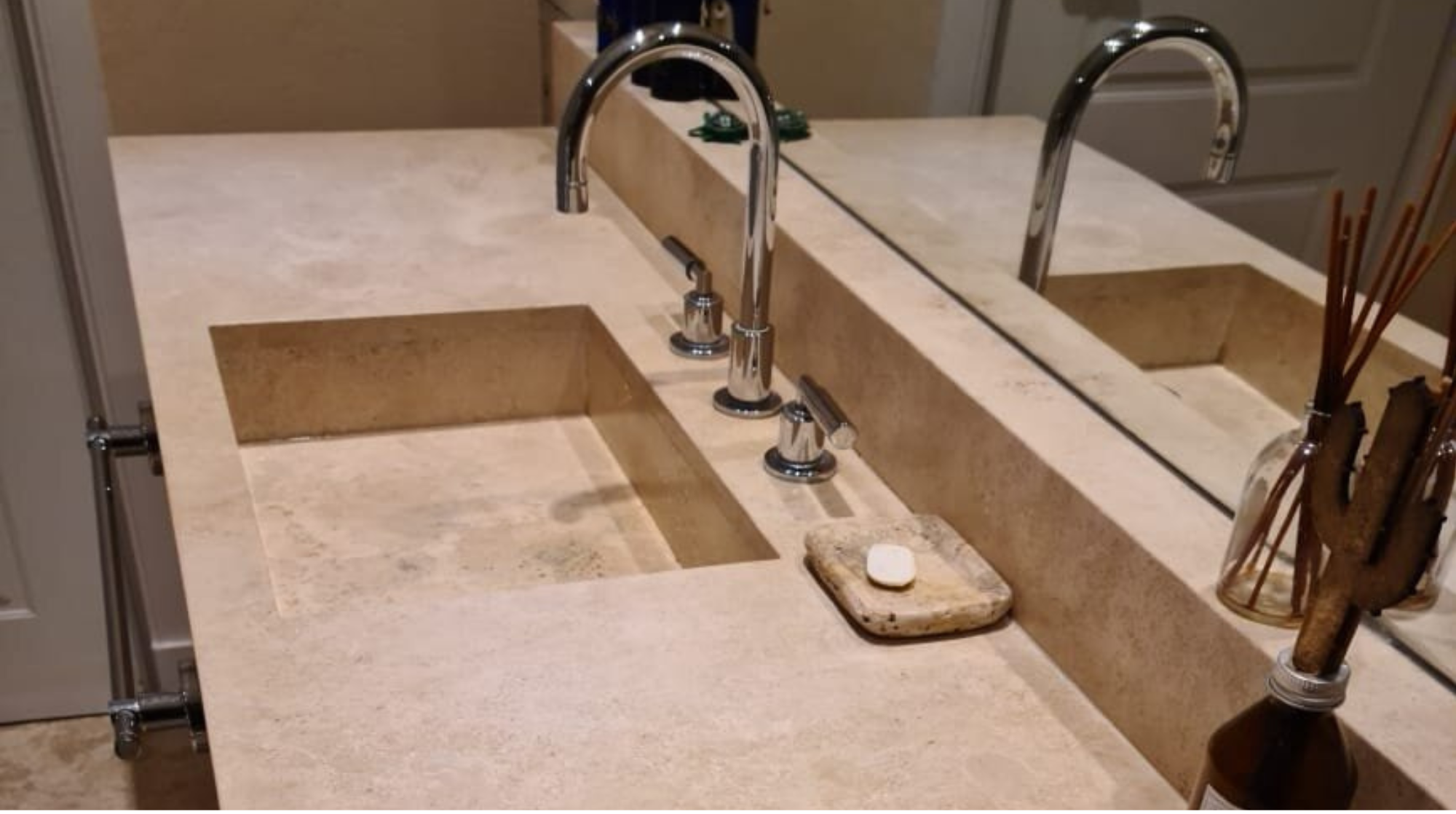 Encimera de cocina de piedra con dos fregaderos, un espejo, y accesorios de cocina en un ambiente doméstico.