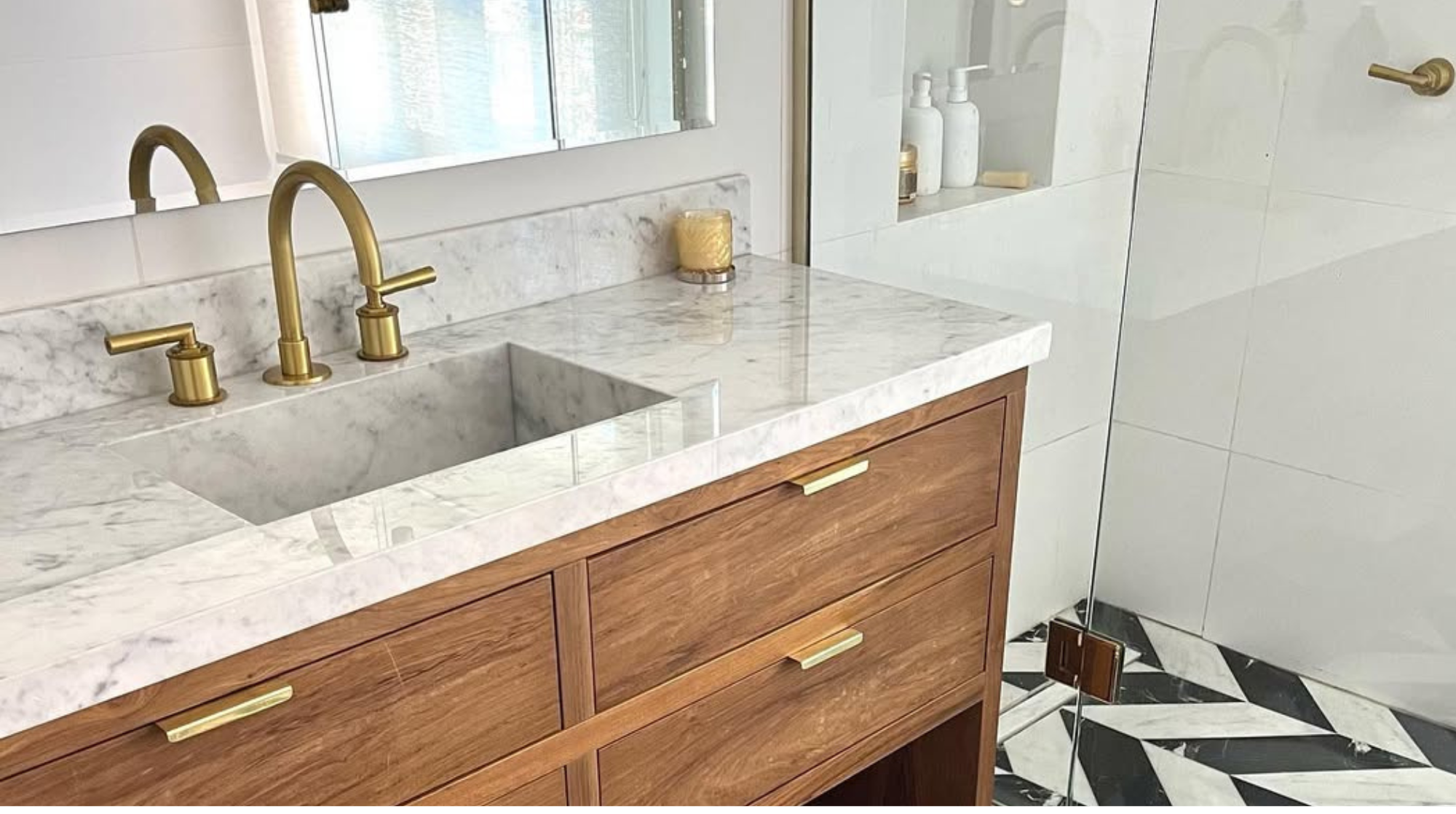 Lavabo de mármol con grifería dorada, espejo en la pared, cerámica blanca y madera en el mueble, y ducha con puerta de vidrio y piso de baldosas negras y blancas.