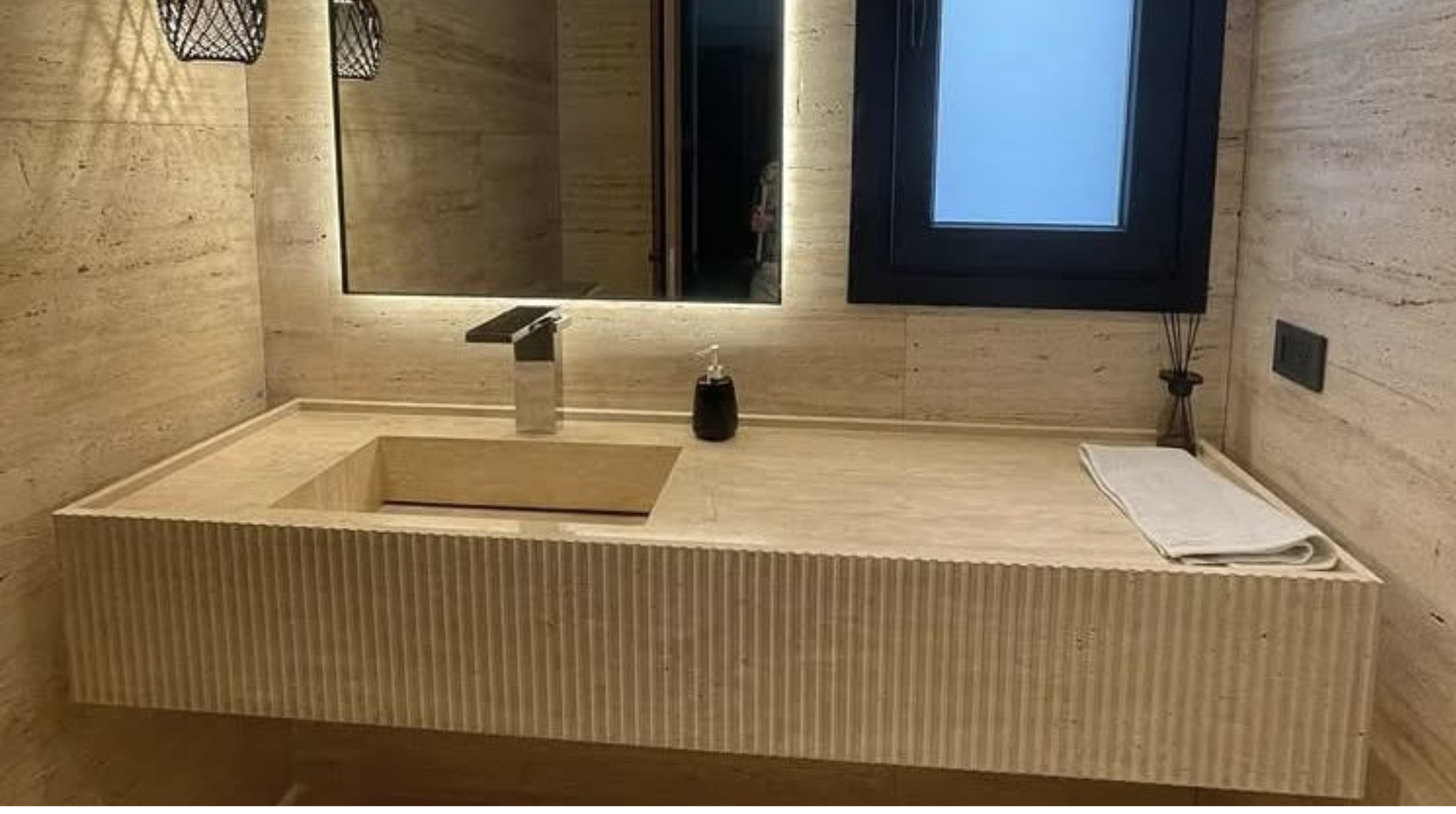 Lavabo de momento en un baño decorado con piedra o mármol, con un grifo moderno, un espejo grande, una ventana pequeña con marco negro, una toalla y un recipiente con difusor de aromas.