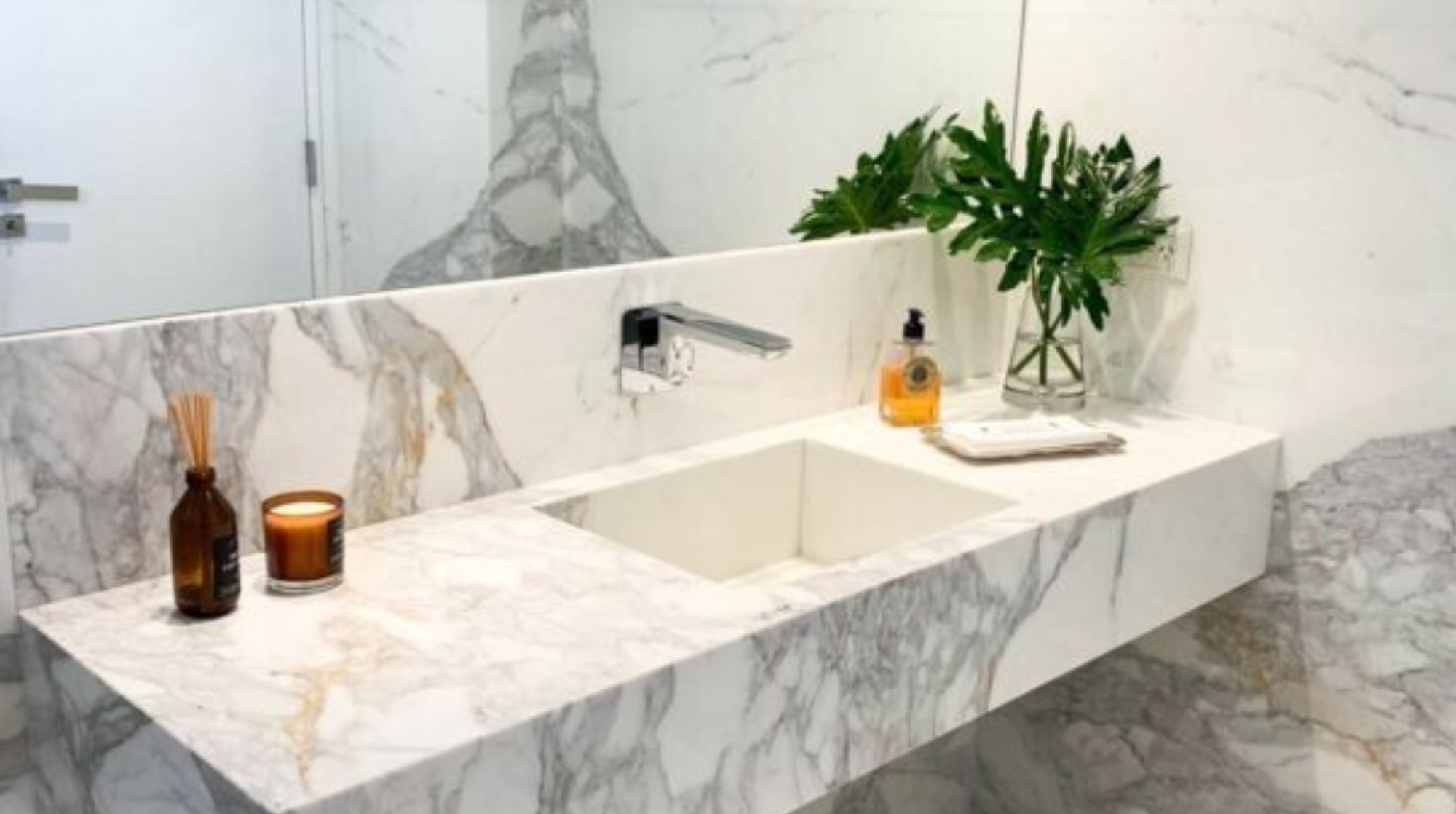 Lavabo de mármol blanco con accesorios decorativos, una planta en un florero y una botella de jabón líquido.