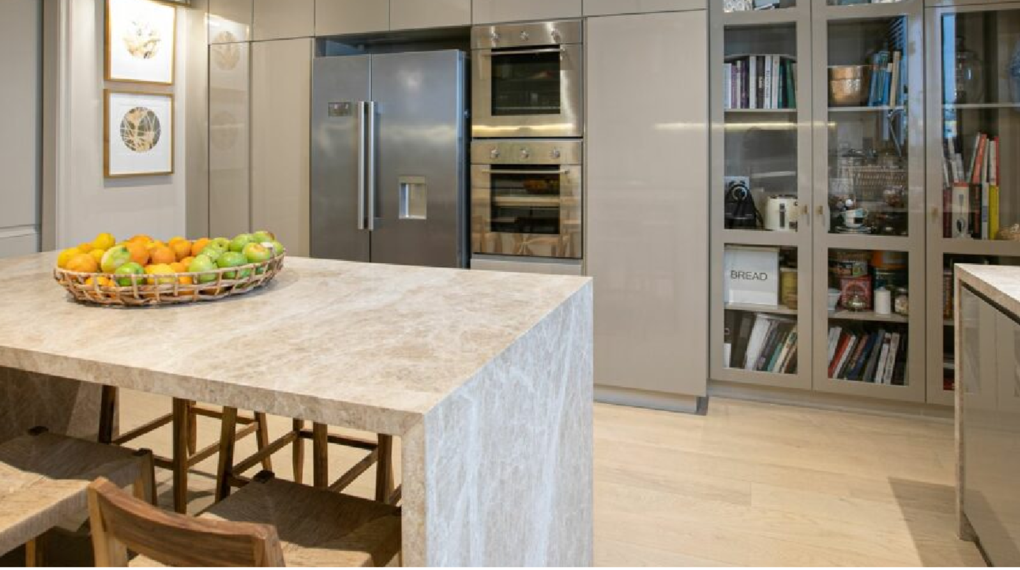 Cocina moderna con isla de granito, armarios grises y electrodomésticos de acero inoxidable. En la isla, una cesta de frutas, y en los gabinetes, libros y alimentos almacenados.