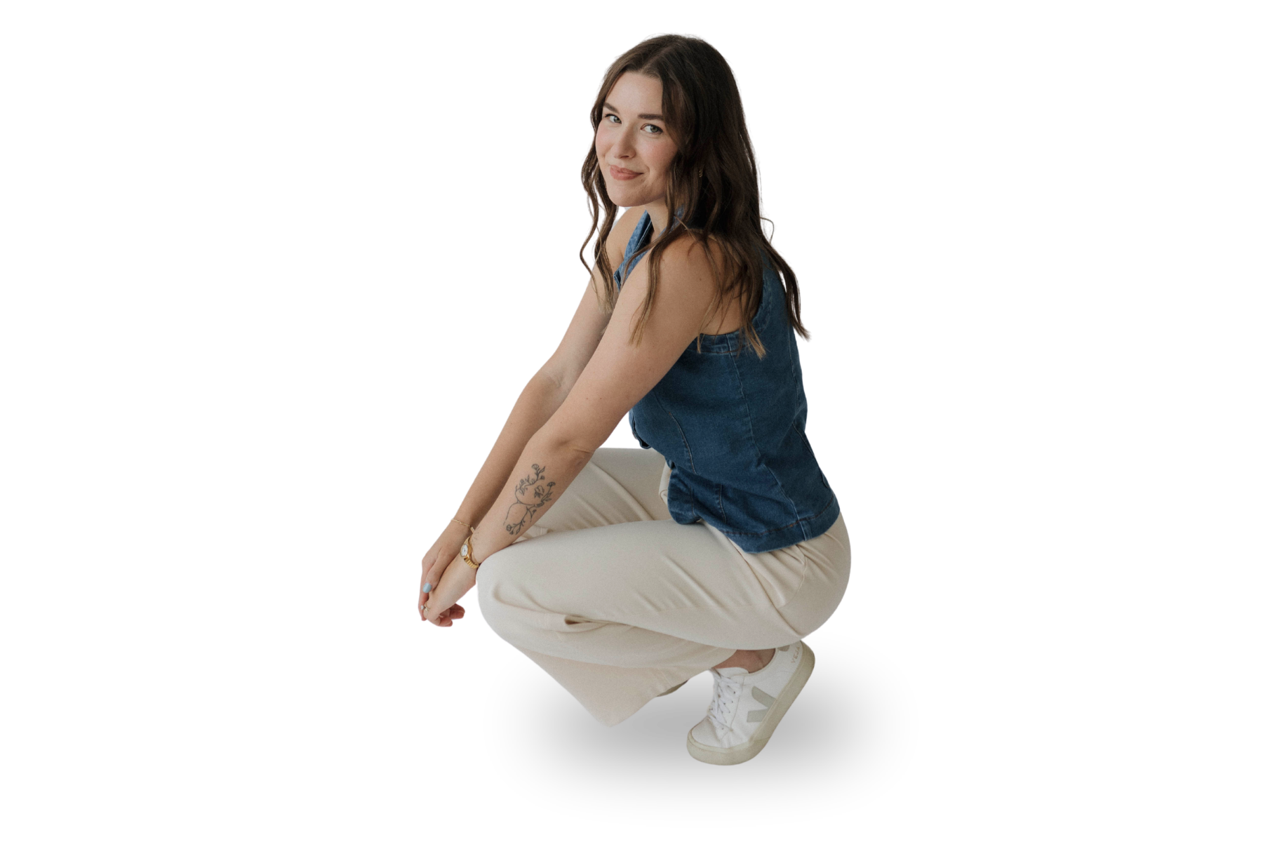 A young woman with long brown hair and a tattoo on her left forearm, wearing a denim sleeveless top, beige pants, and white sneakers, is squatting with her knees bent, looking at the camera with a slight smile.