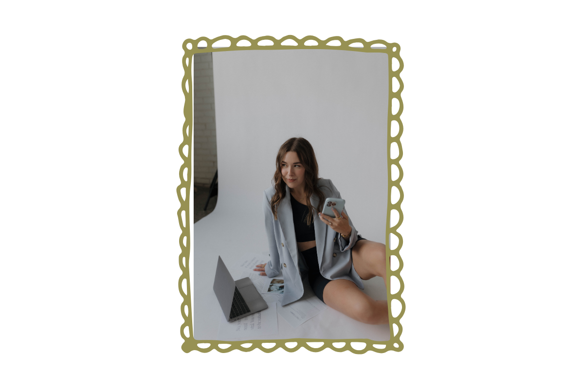 Young woman sitting on the floor in front of a white background, wearing a gray blazer and black clothing, holding a smartphone, with a laptop and papers around her.