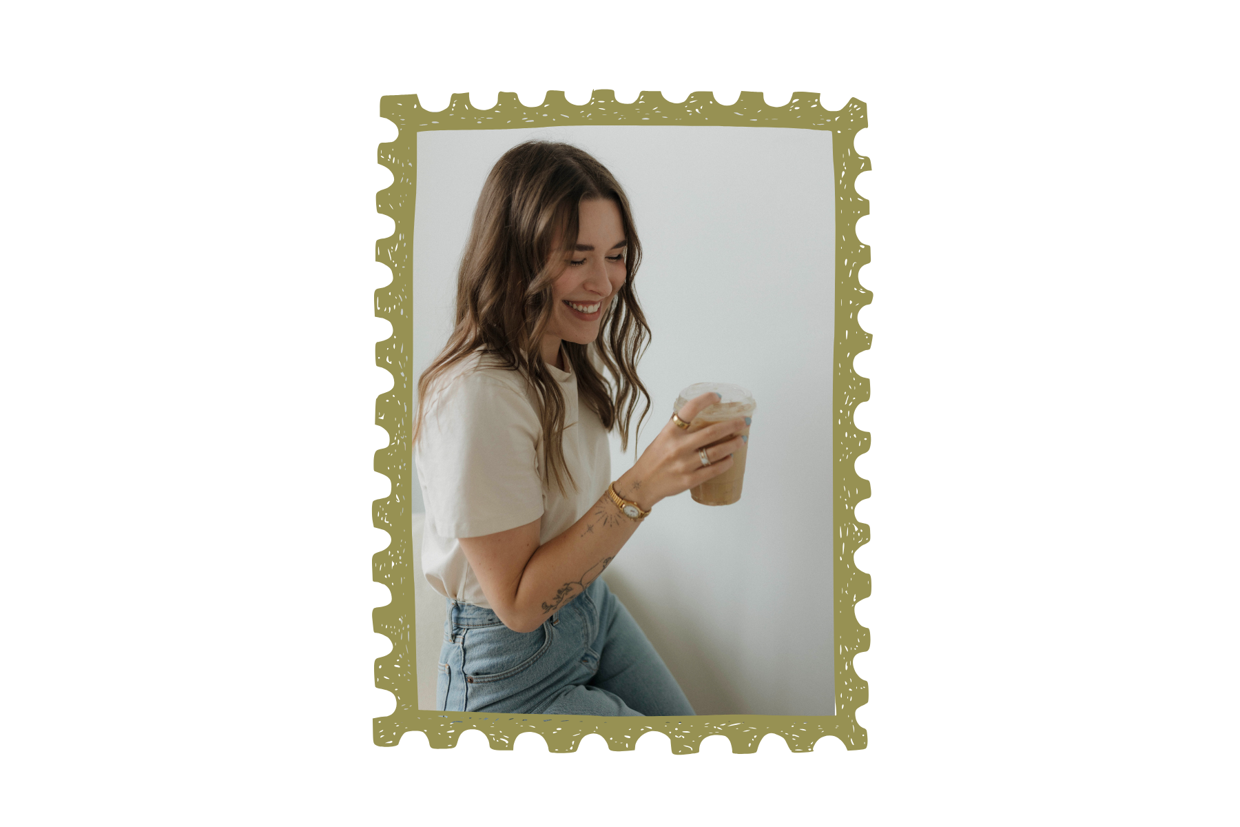 A young woman with wavy brown hair is smiling and looking down at her iced coffee in a clear plastic cup. She is wearing a beige t-shirt, high-waisted jeans, and gold jewelry, and has tattoos on her arm. She is sitting against a white background.