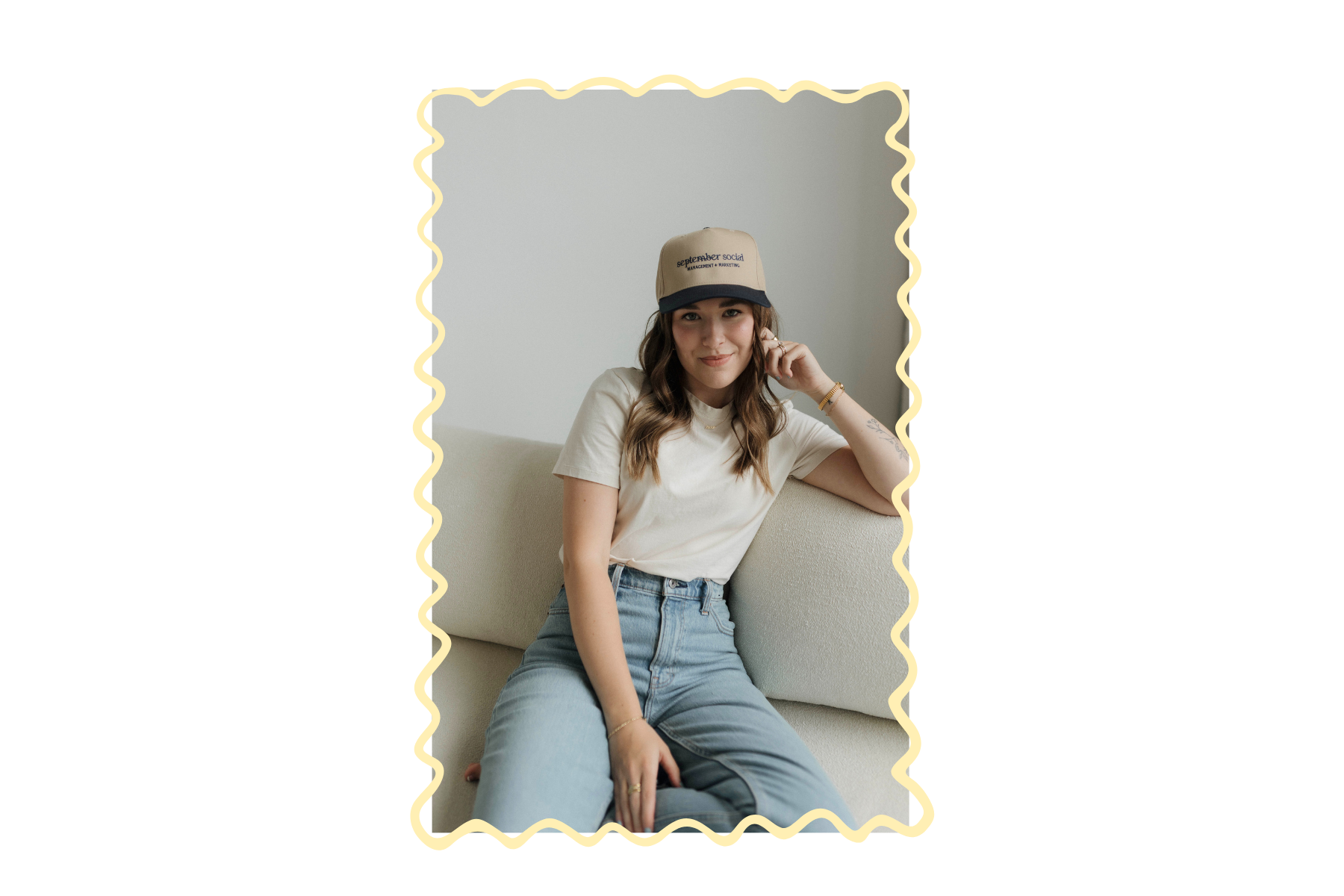 Young woman sitting on a light-colored sofa, wearing a beige cap, white t-shirt, and jeans, smiling and looking at the camera.