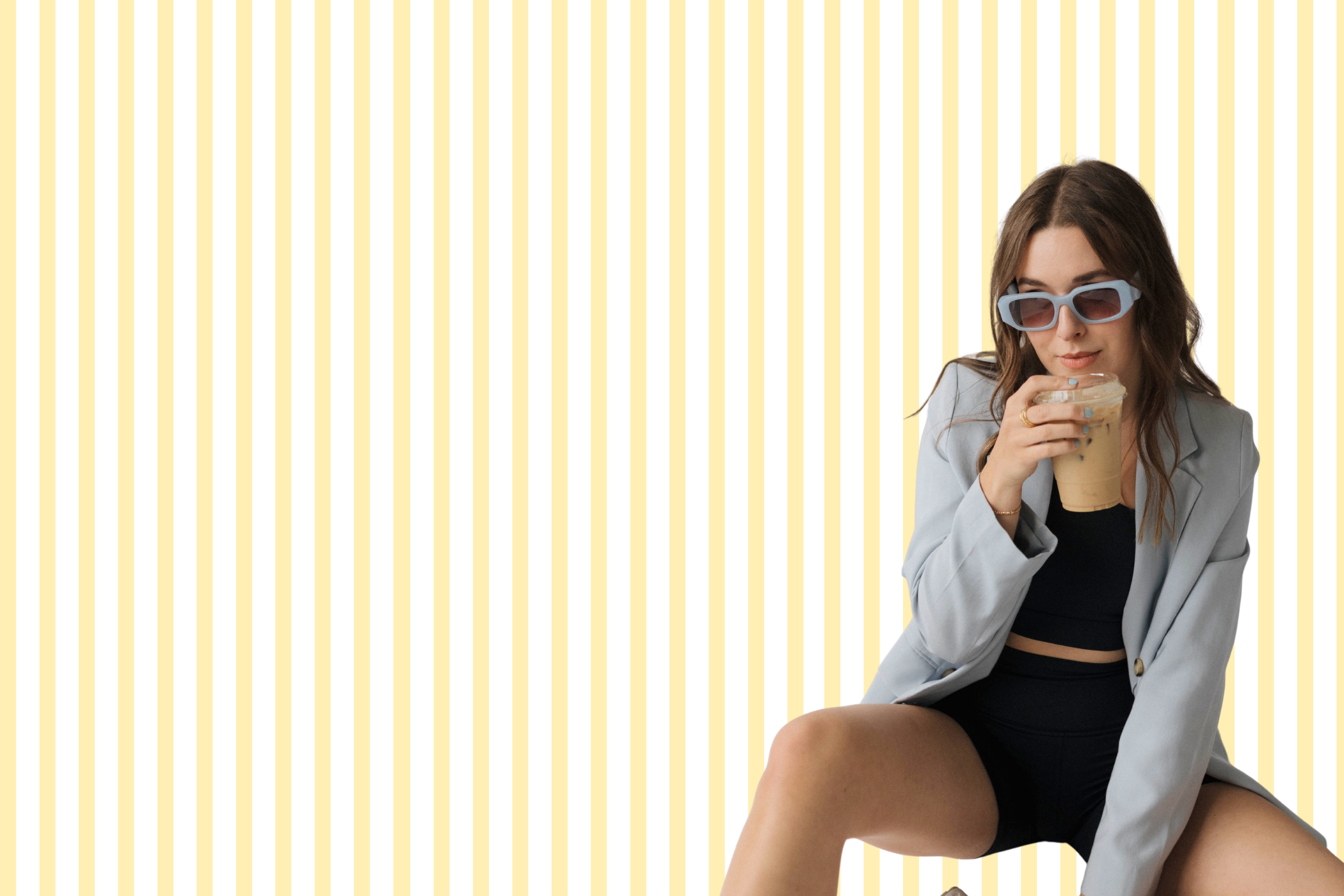A woman with long brown hair wearing large sunglasses and a gray blazer, holding a coffee drink with black straws, seated with her legs crossed against a pale yellow background with vertical stripes.