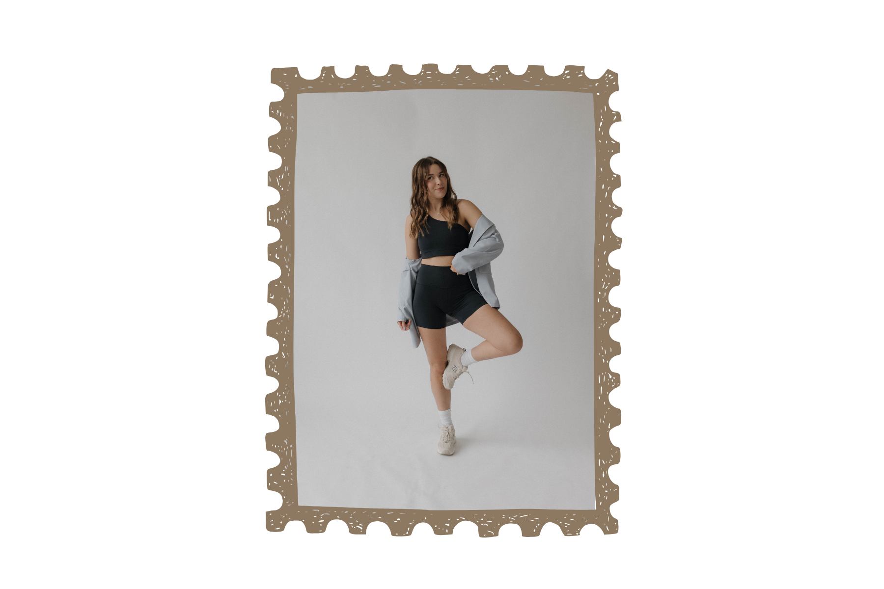 Young woman in black sports bra and shorts posing with one leg raised, wearing an oversized gray jacket, white socks, and sneakers against a plain white background.