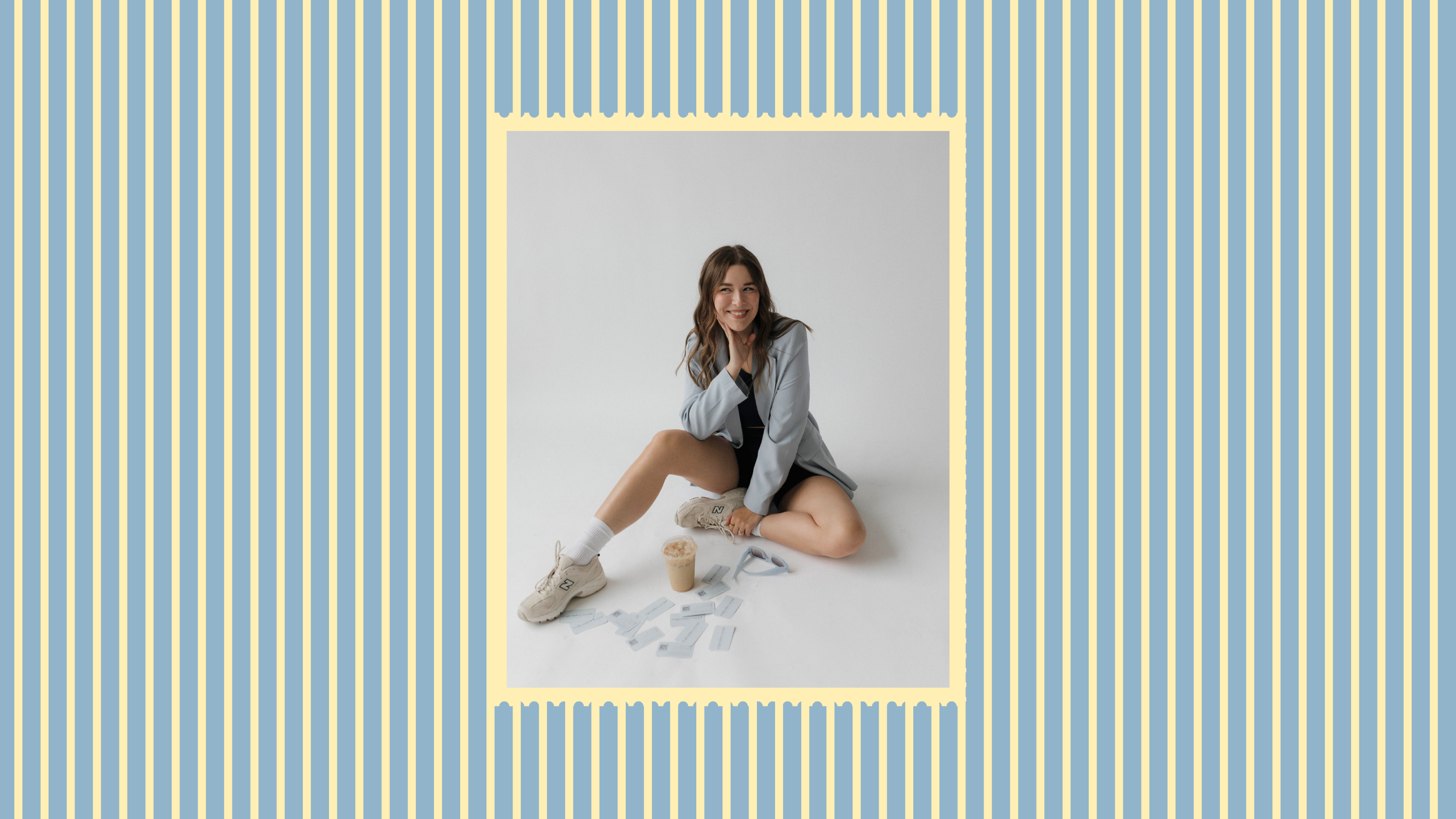 A woman sitting on the floor surrounded by scattered cards, a coffee drink, sneakers, and sunglasses, smiling and posing with one hand on her face.
