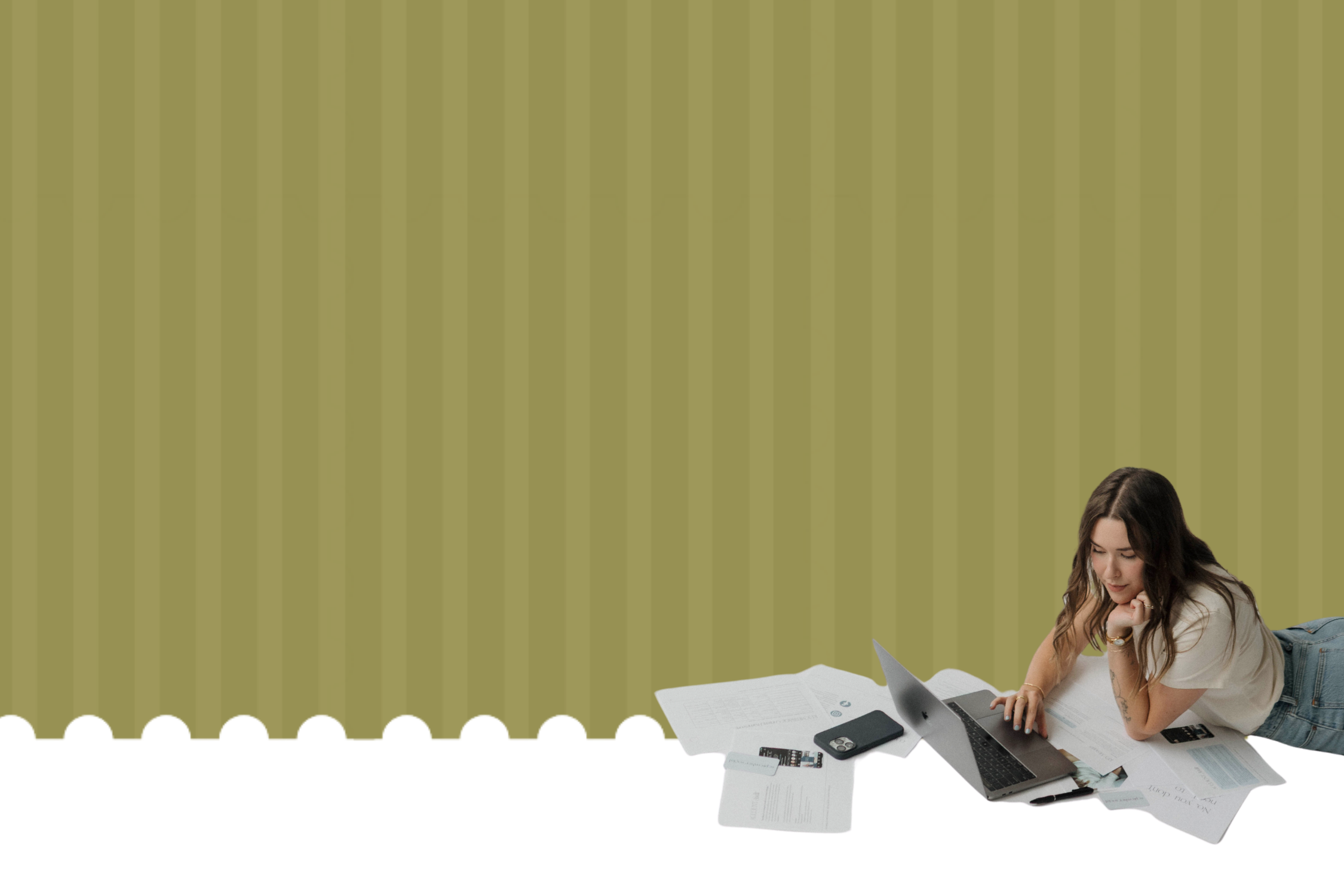 A woman with long wavy hair lying on her stomach on a cluttered desk, working on a laptop. The desk has various papers, a smartphone, a calculator, and other office supplies, with a plain green wall in the background.