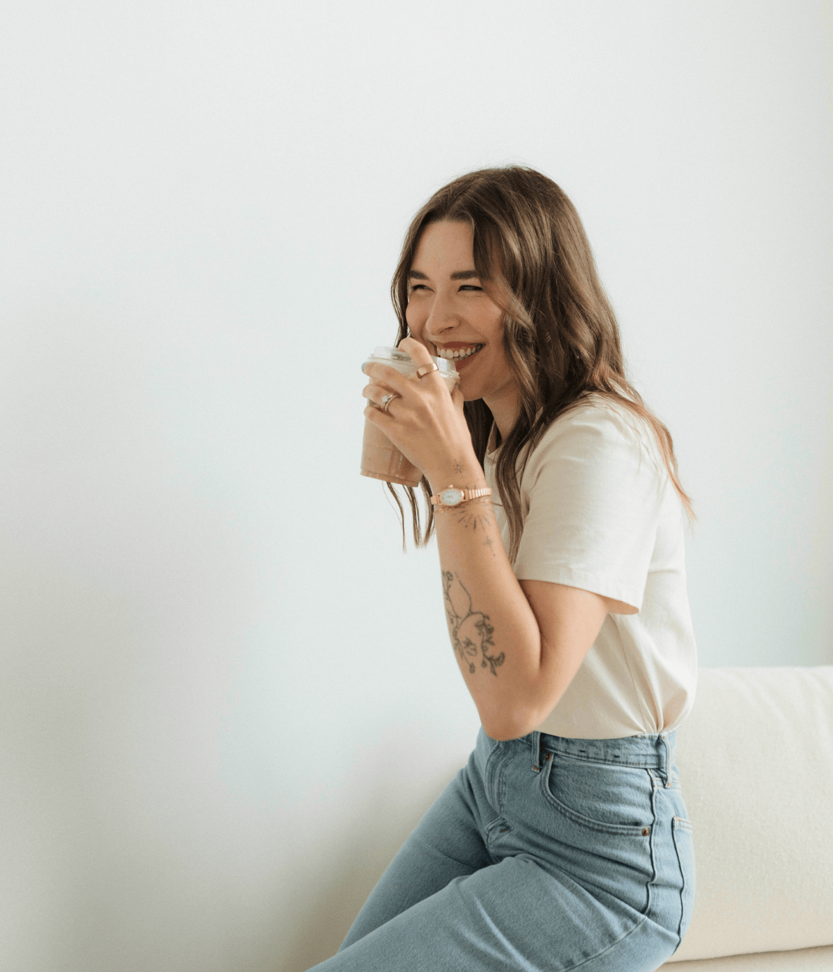 A young woman with tattoos and wavy brown hair sitting on a beige sofa, smiling and drinking a coffee in a white t-shirt and light blue jeans against a plain white wall.