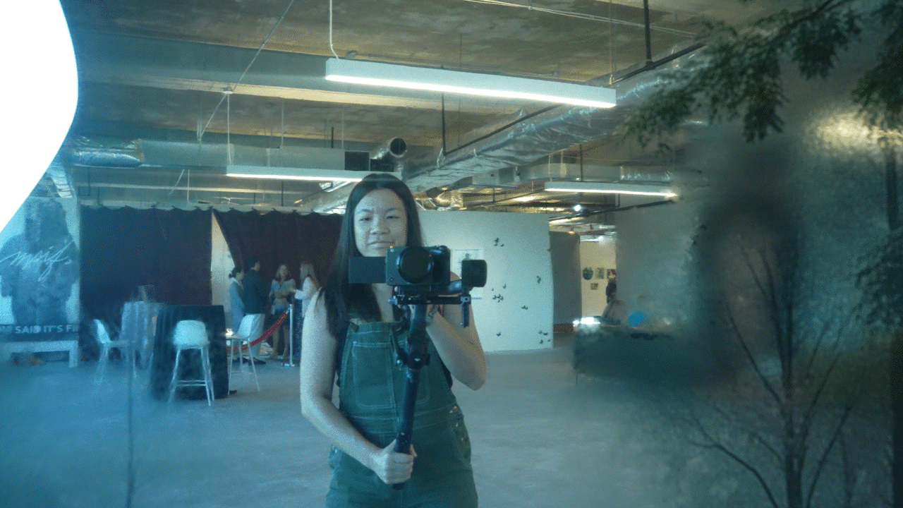 A woman taking a selfie in front of a mirror with a camera mounted on a tripod in an indoor space with industrial ceiling ductwork and a group of people in the background.