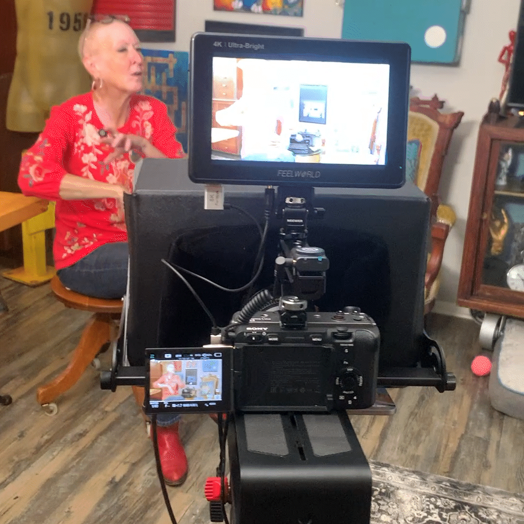 A woman in a red floral blouse and dark pants seated on a wooden chair, being filmed in a room with various framed artworks and a yellow chair in the background, with a camera setup in the foreground.