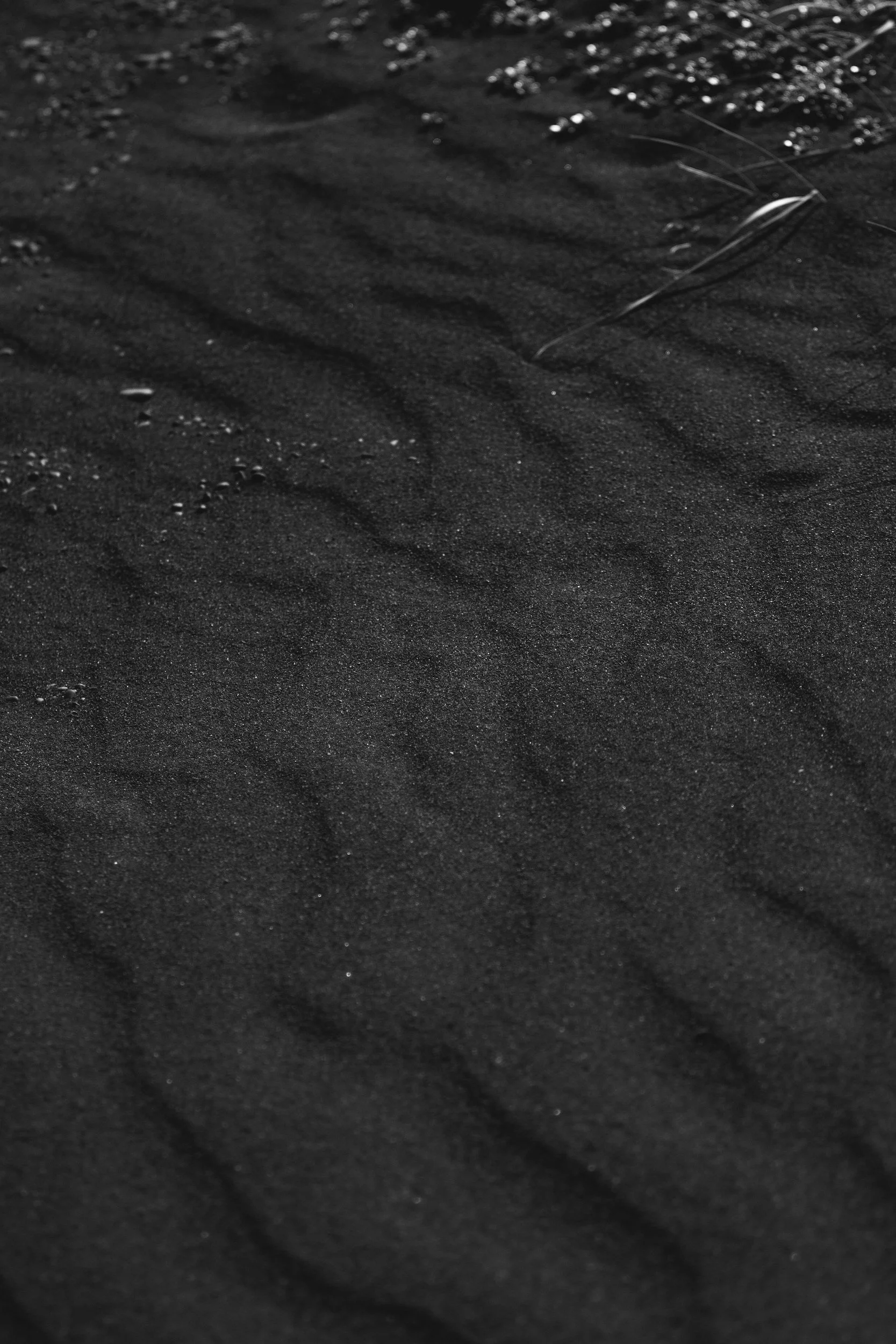 Close-up of black sand with small pebbles and a slightly bent piece of grass.