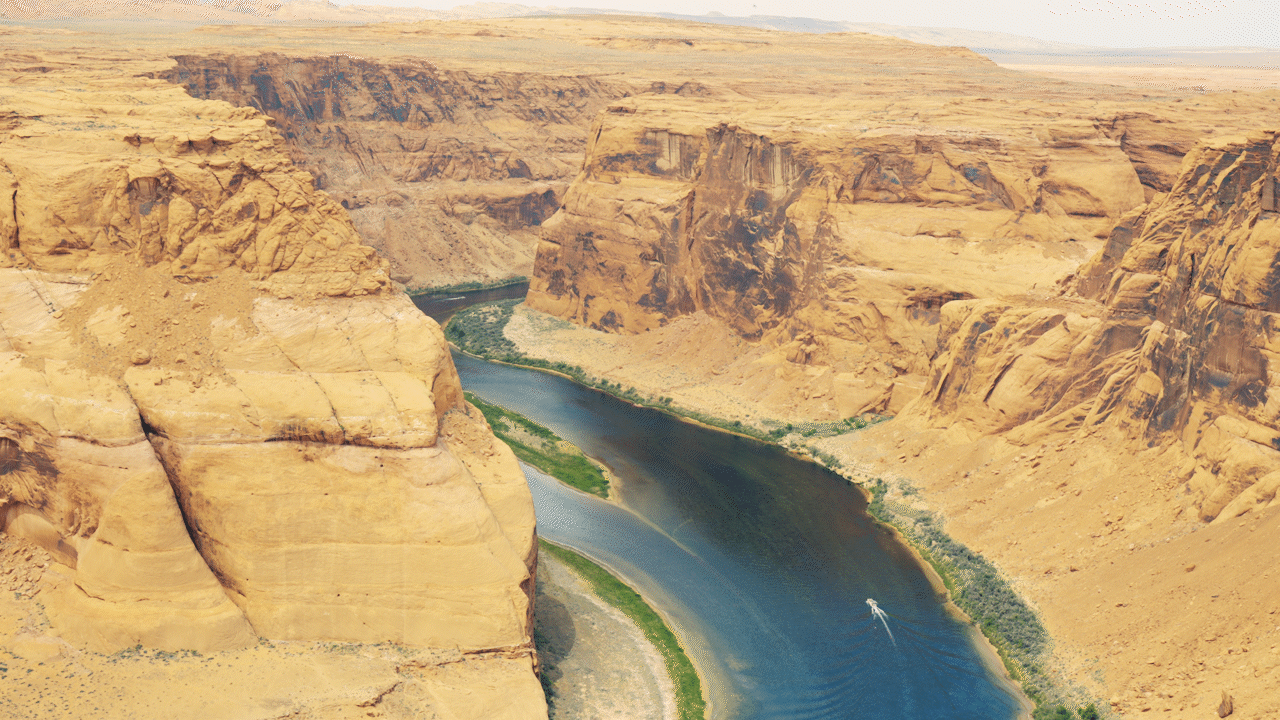 Aerial view of the Grand Canyon with a winding Colorado River at the bottom, surrounded by steep, layered desert cliffs.