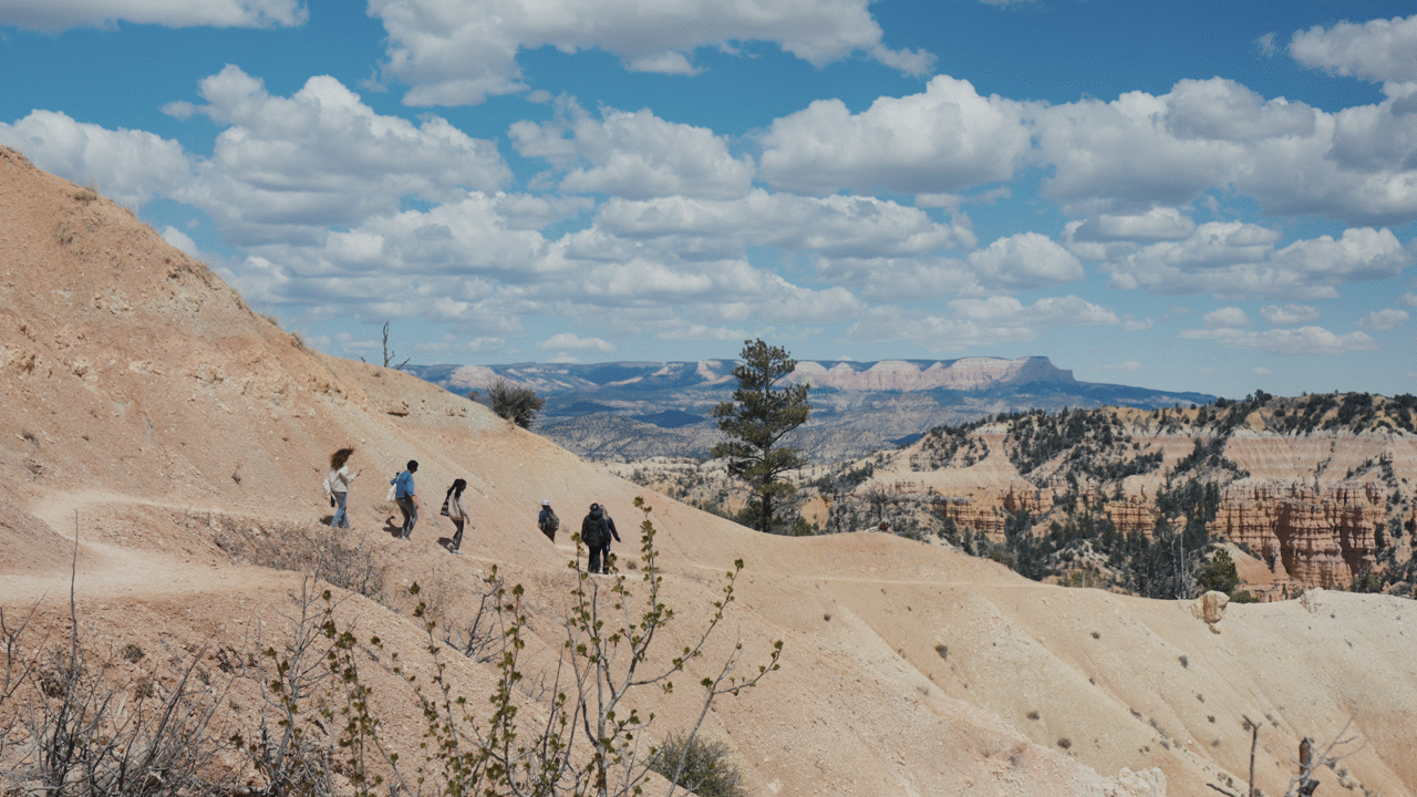 A group of six people hiking on a dirt trail in a desert landscape with layered rock formations under a partly cloudy sky.