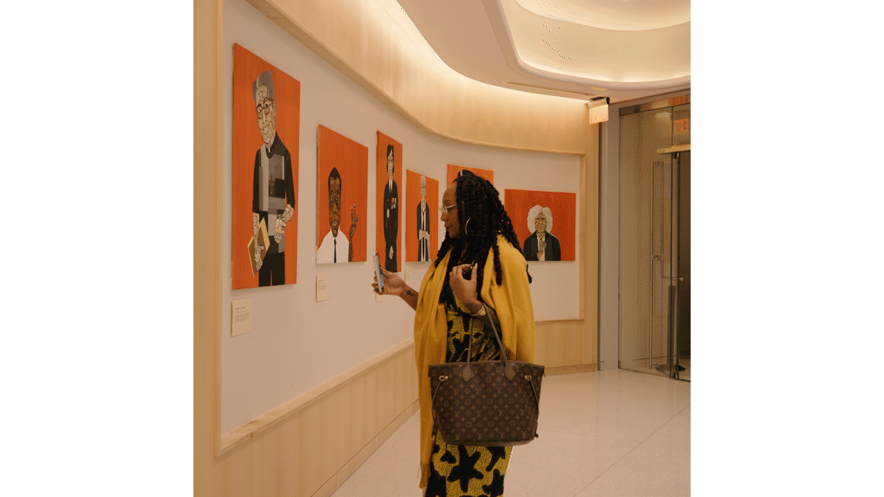 A woman with glasses and braided hair wearing a yellow coat and black-and-yellow patterned dress standing in an art gallery, looking at colorful portraits of elderly people on the wall.