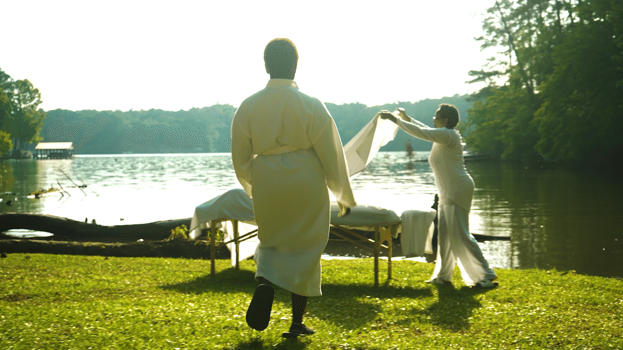 Two women outdoors near a lake, one in a long white robe walking toward the water, the other in a long white dress holding a cloth, with a massage table covered with white fabric nearby and trees in the background.