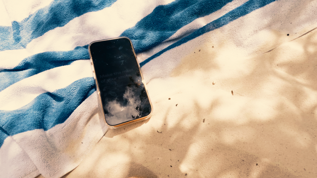 A smartphone lying on a beach towel, with the towel partially in the sand. The towel is blue and white striped, and the phone's screen reflects a cloudy sky.