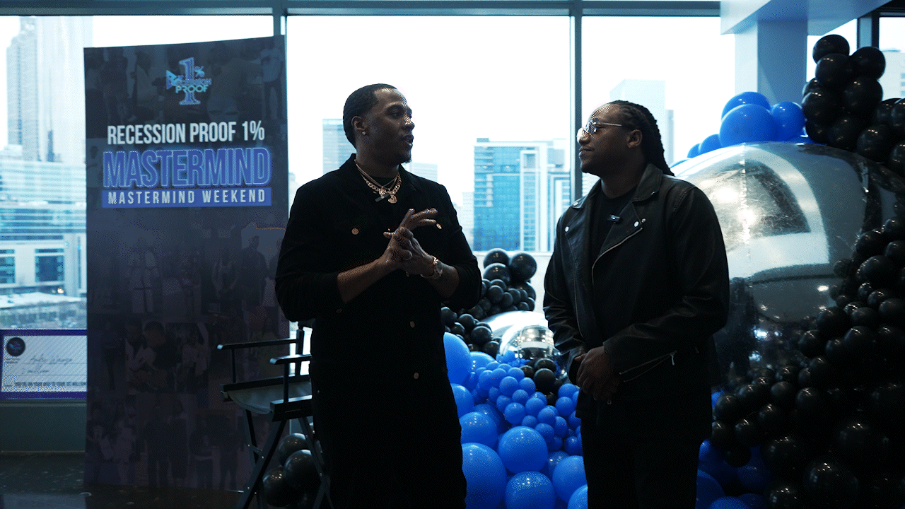 Two people having a conversation in an indoor setting with large windows, city buildings outside, black and blue balloon decorations, and a sign that reads "Recession Proof 1% Mastermind Weekend."