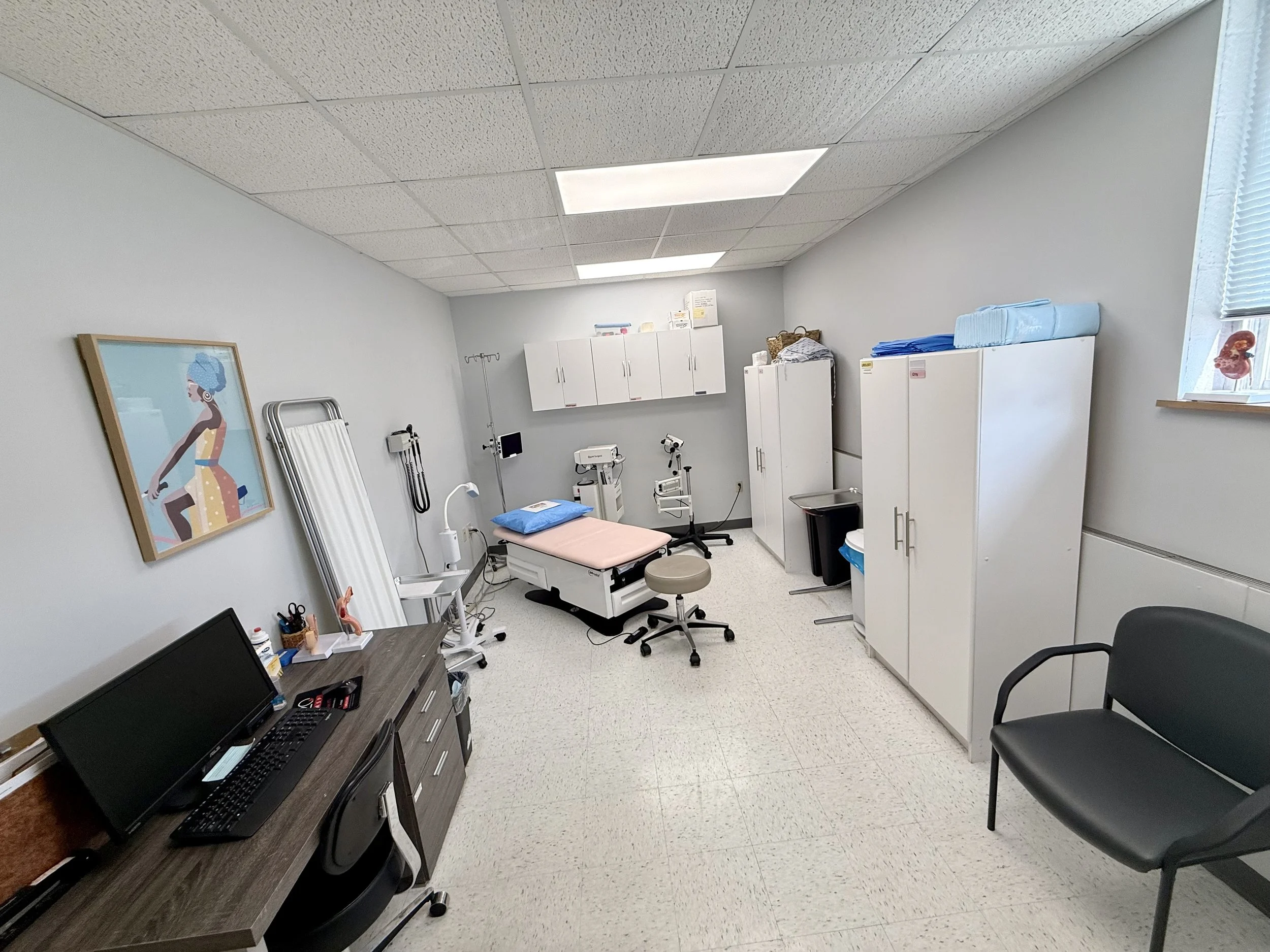 A medical examination room with white walls, ceiling tiles, and various medical equipment including a bed, cabinets, and a chair.