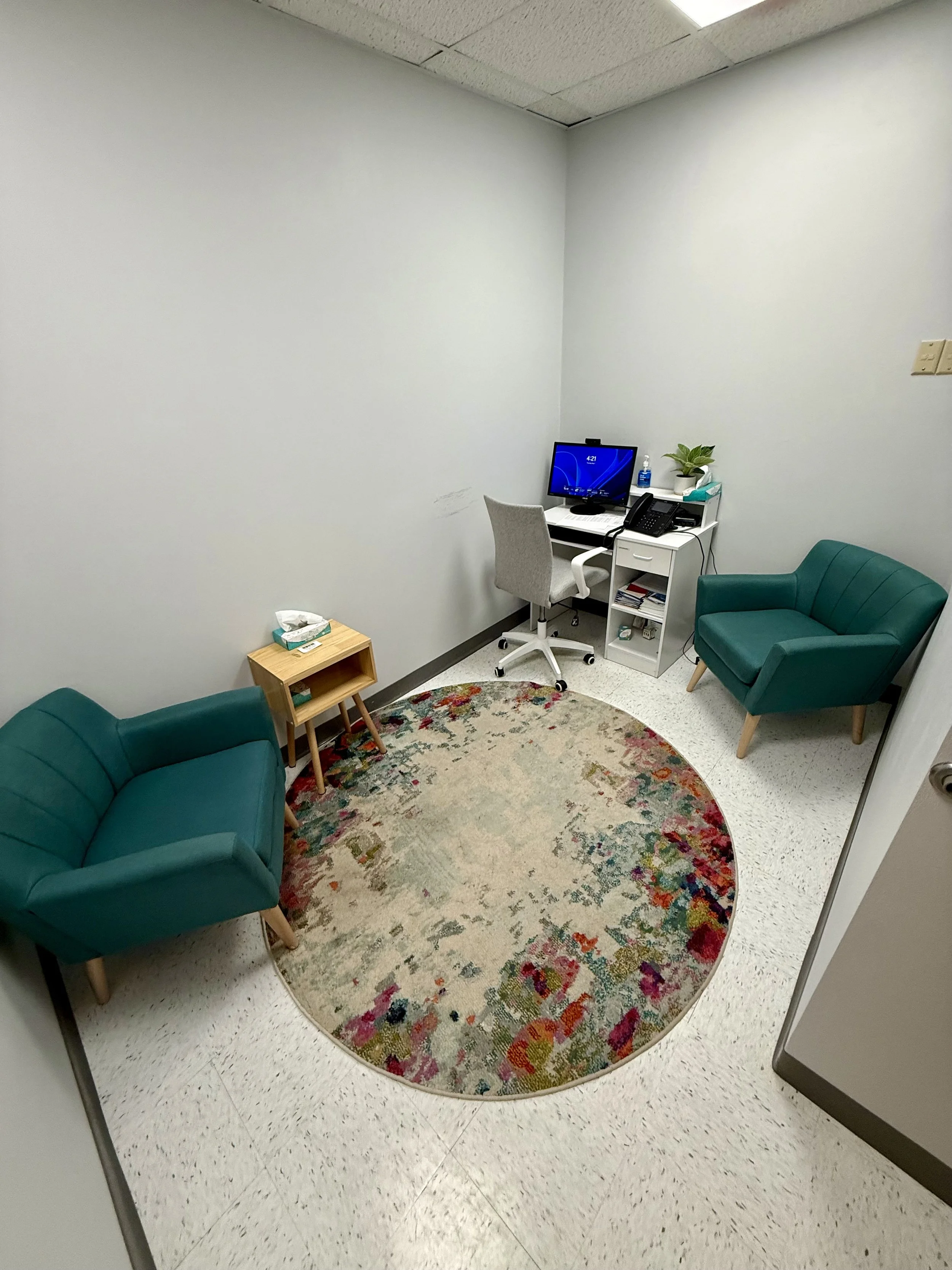 A waiting room with two teal armchairs, a floral oval rug, a small wooden side table with tissue box, a white desk with a computer monitor, phone, hand sanitizer, and potted plant, against plain white walls and a ceiling with fluorescent lights.