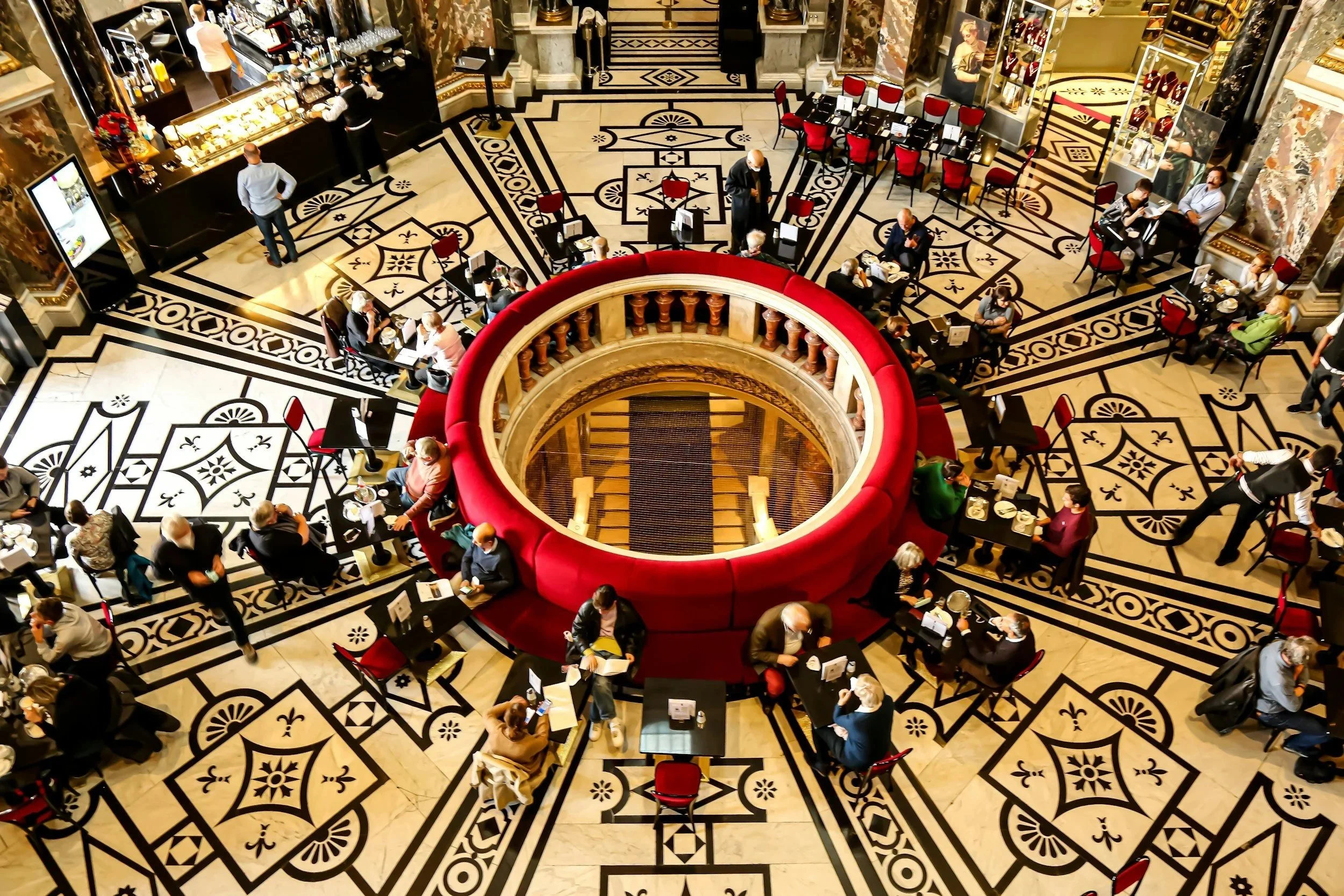 A bustling cafe in a grand, ornate interior with a circular red cushioned seating area in the center, surrounded by tables and chairs with people dining and socializing. The patterned marble floor adds elegance to the scene.
