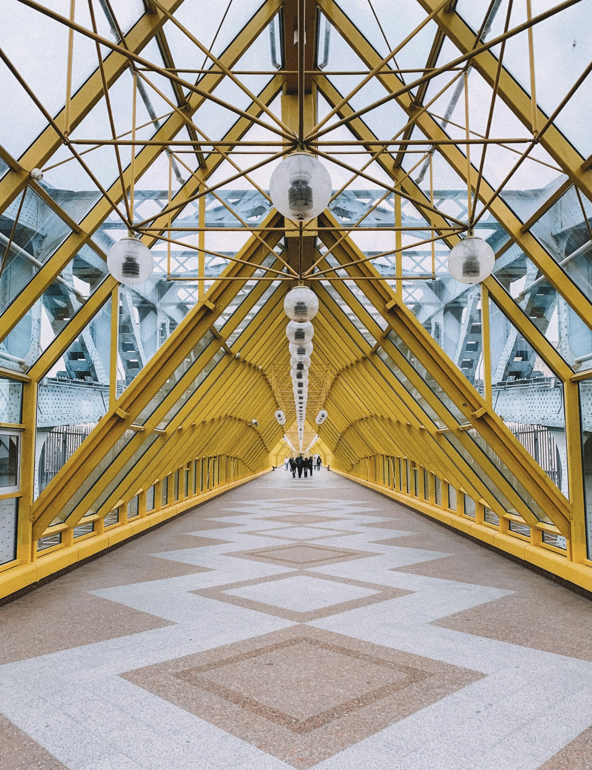 A covered pedestrian bridge with yellow metal framing and glass panels, illuminated by round ceiling lights, with people walking in the distance.