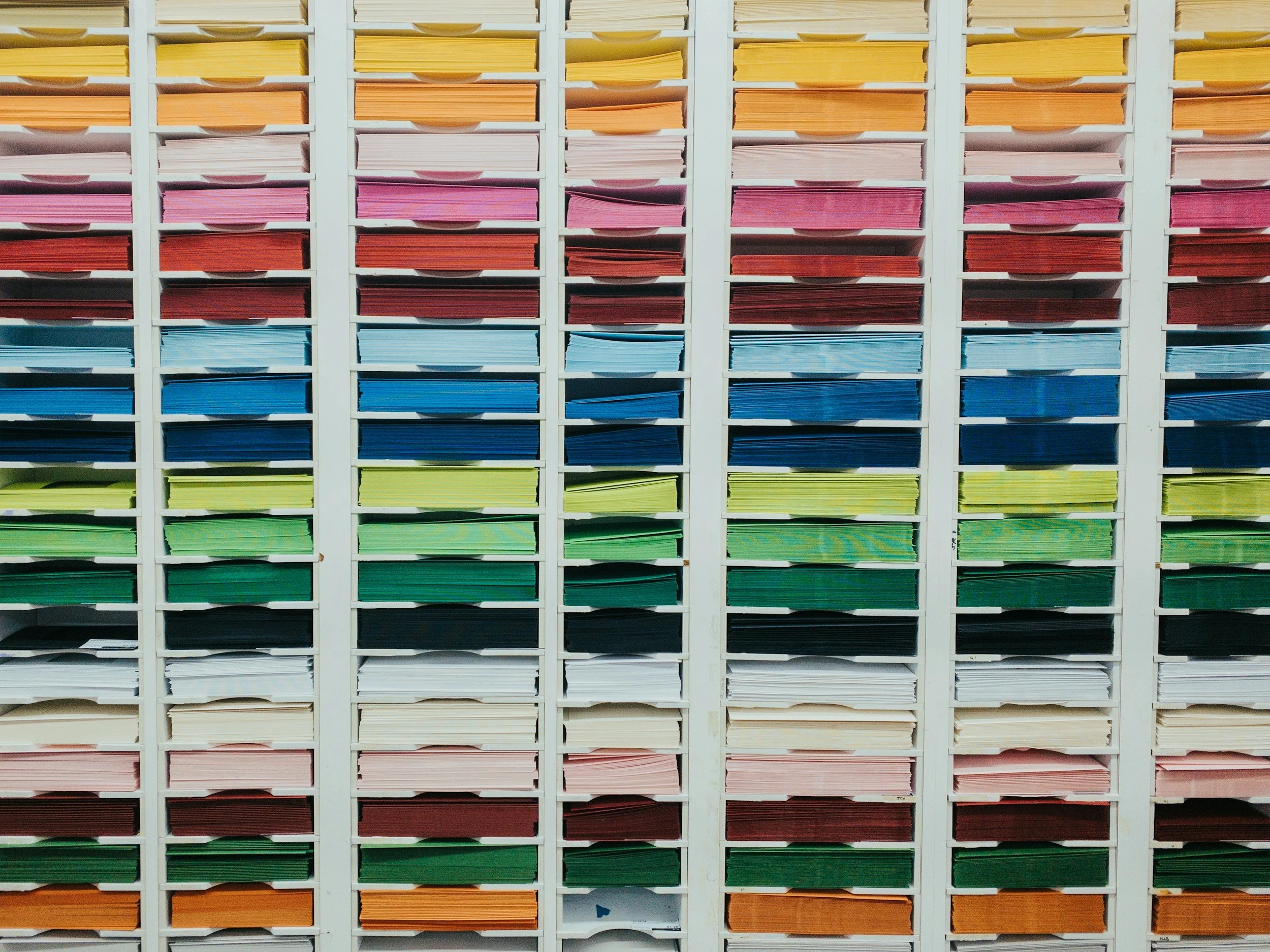 Color-coded paper folders arranged in a vertical filing cabinet with multiple shelves, organized by color in a gradient order.