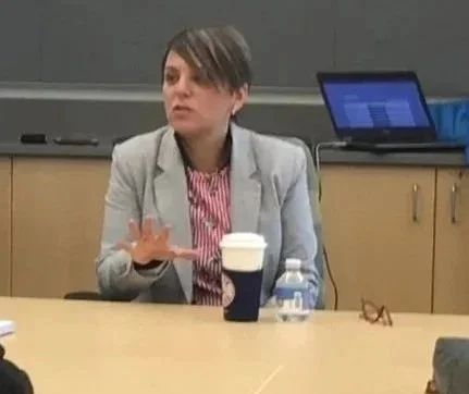 Stacey Weissbock in a gray blazer sitting at a conference table with a coffee cup, water bottle, and glasses in front of her, during a meeting.