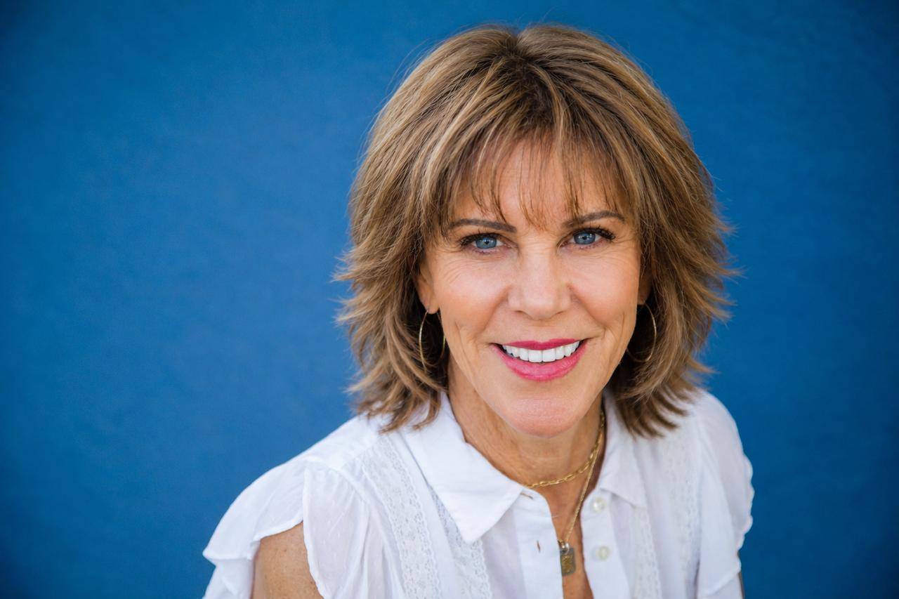A smiling middle-aged woman with short, layered brown hair with highlights, wearing a white collared blouse, gold jewelry, and earrings, against a blue background.