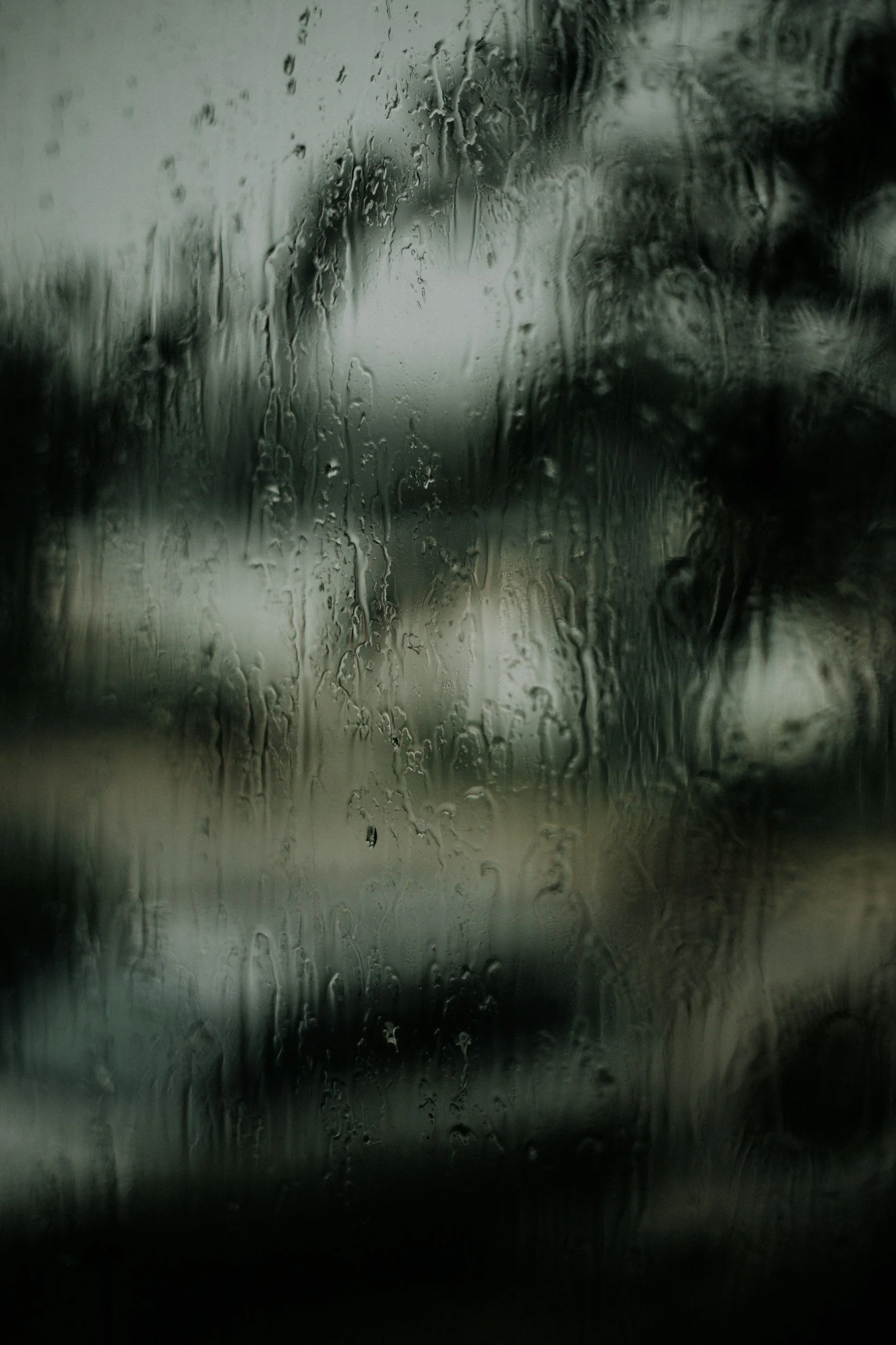 Rainy window with water droplets and streaks, obscuring the view of a blurred outdoor scene.