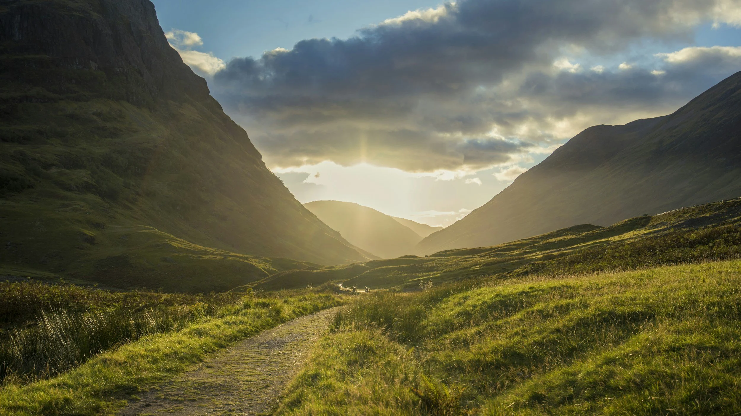 Sunset over a mountainous valley with a rugged dirt path winding through green grass and hills.