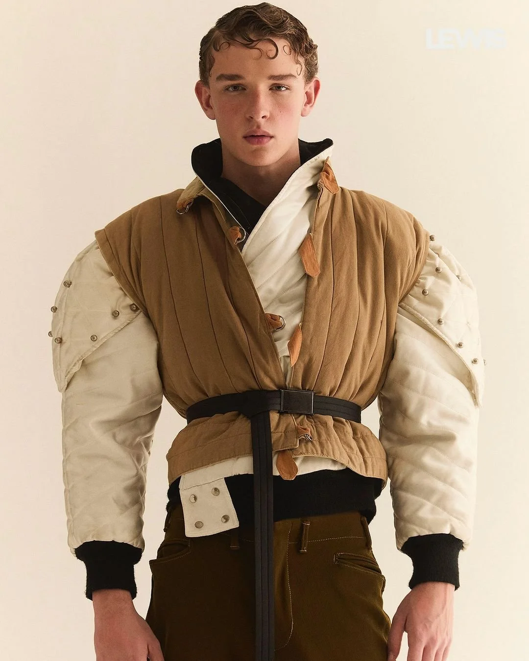 Jovem em um traje inspirado em personagem de ficção científica, com jaqueta de cores caramelo e branco, calças marrom e fundo plain.