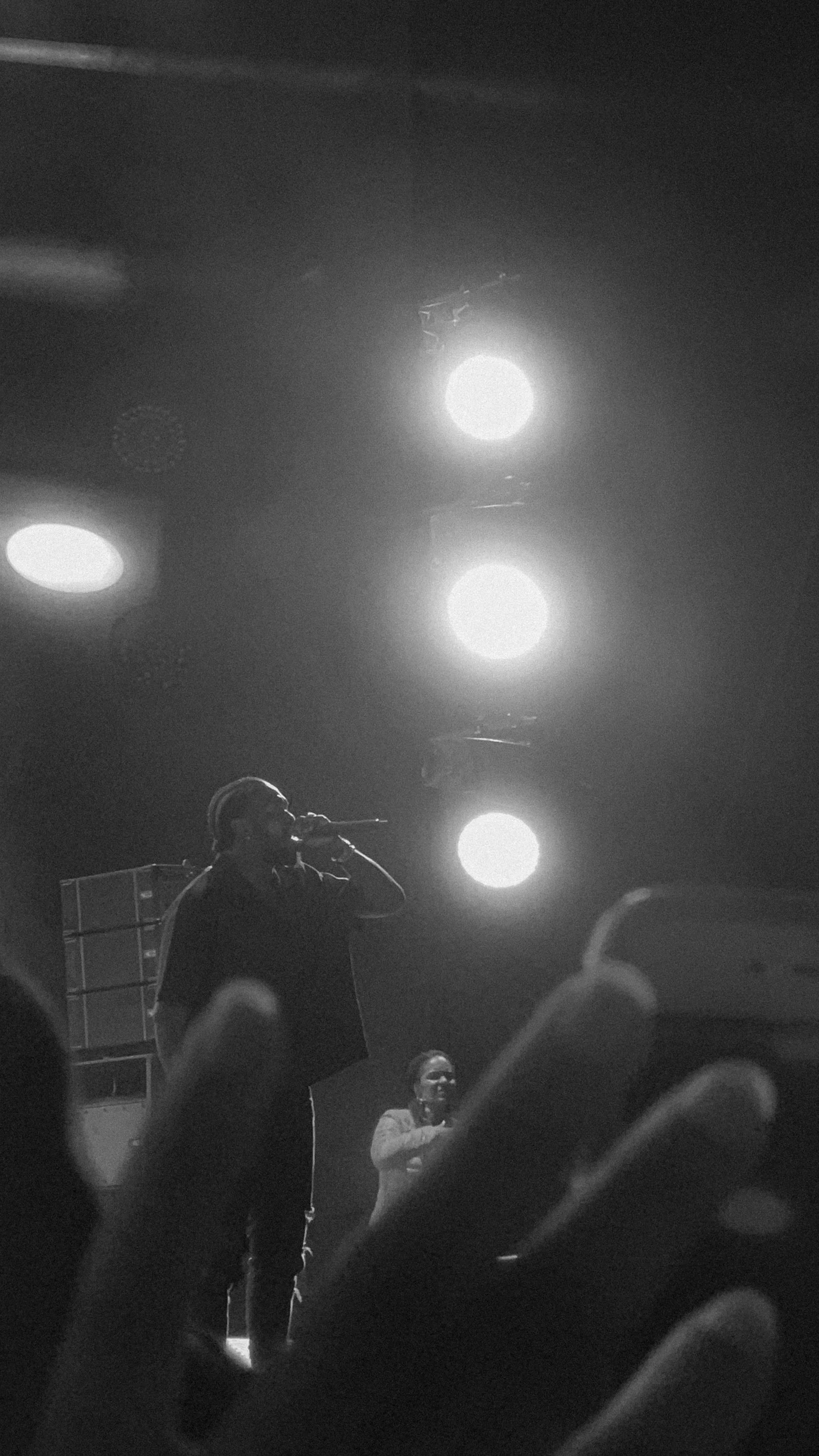 Performers on stage during a concert, with bright stage lights overhead, and an audience in the foreground.
