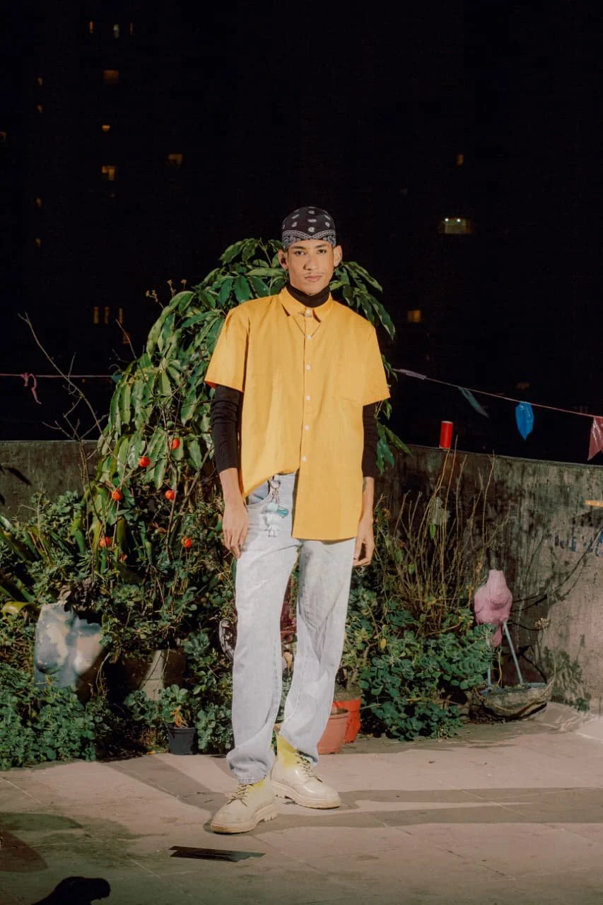 Pessoa jovem posando ao ar livre à noite, vestindo uma camiseta amarela, calça jeans e boné, com plantas ao fundo.