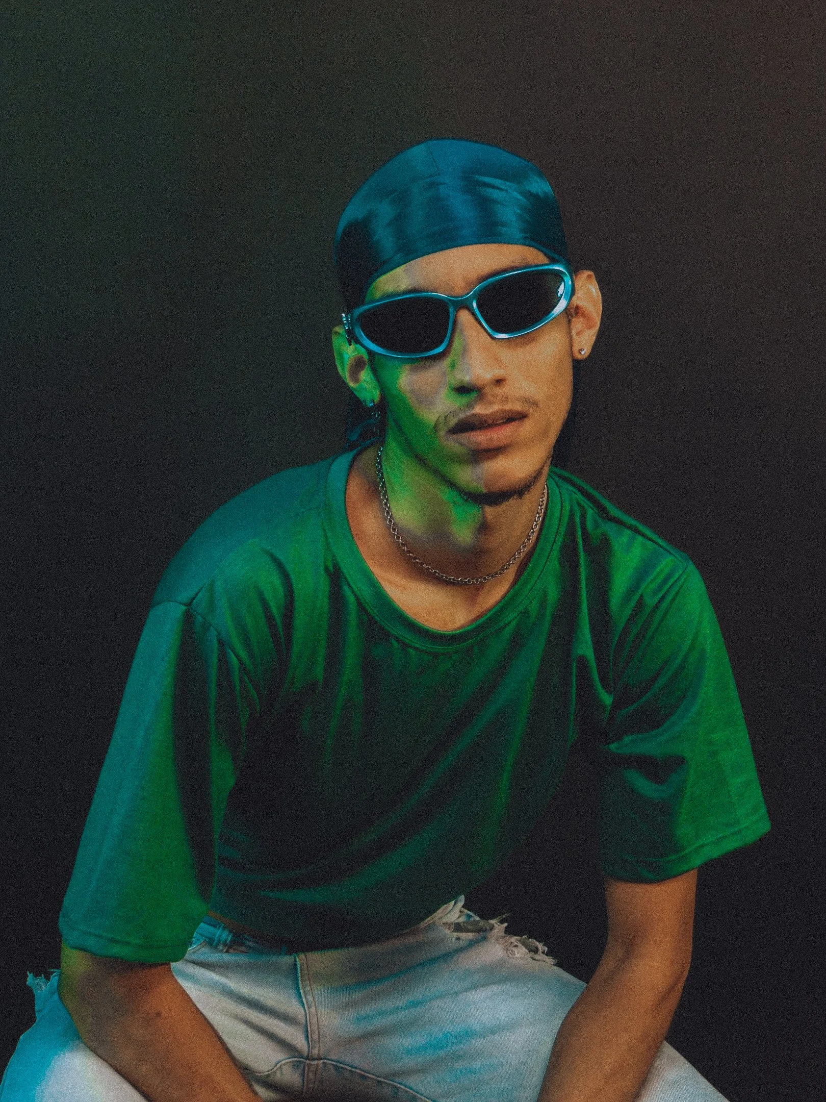 Pessoa usando camiseta verde, óculos escuros azuis, bandana azul, com fundo escuro.