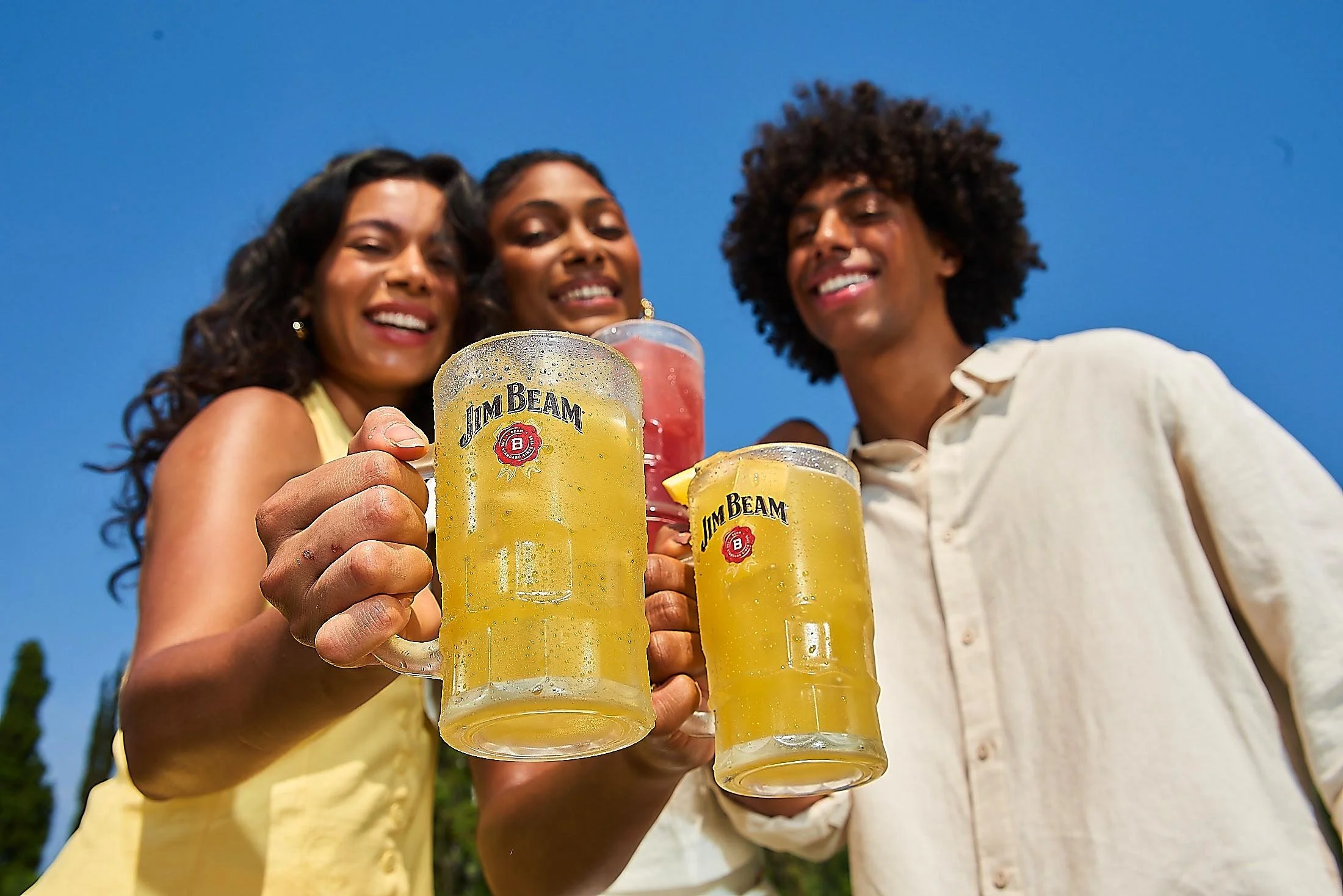 Três pessoas sorrindo e brindando com canecas de cerveja Gelada Jim Beam, ao ar livre com céu azul e árvores ao fundo.