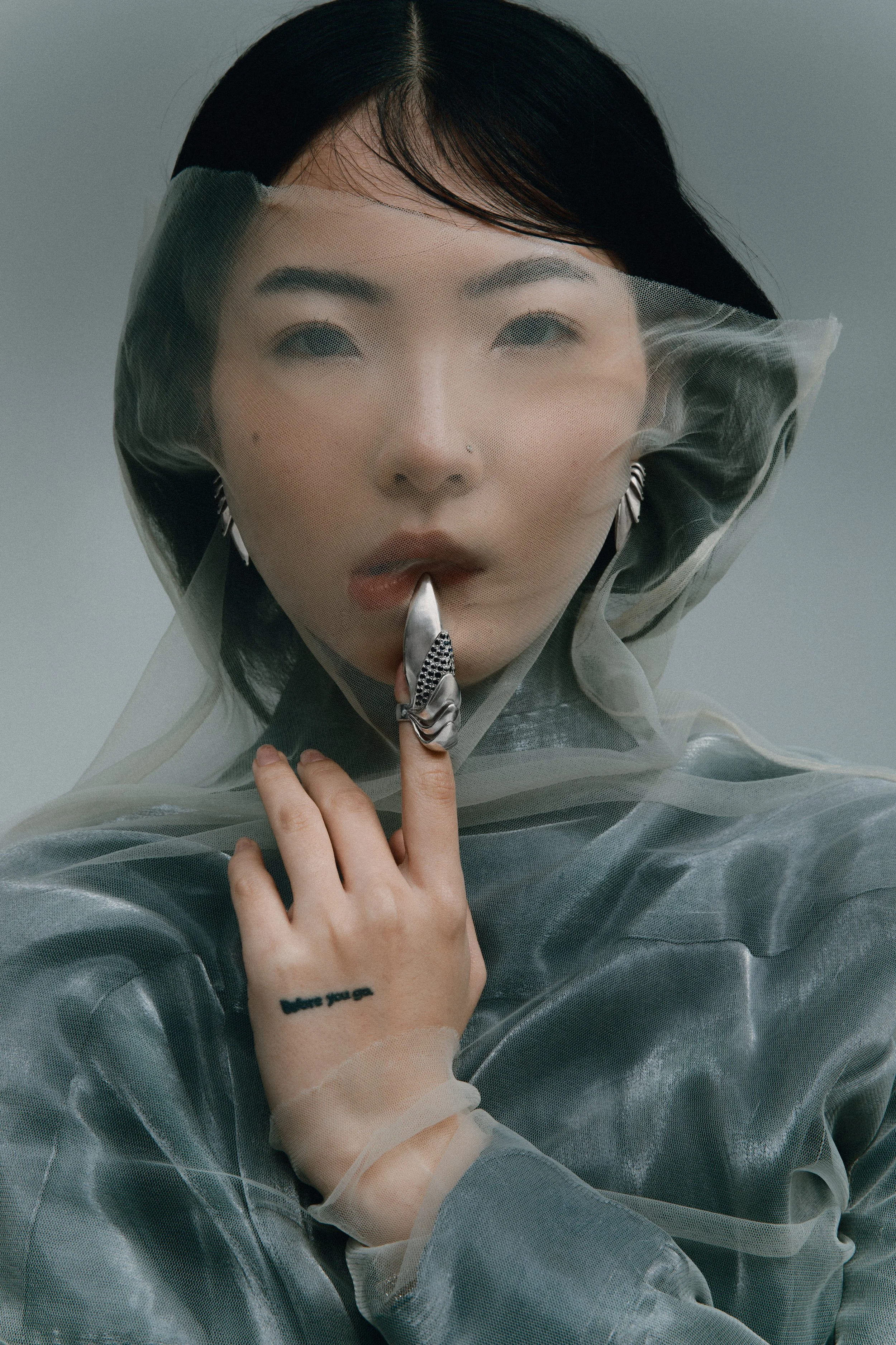 Retrato de mulher com cabelos lisos e escuros, usando uma roupa de tecido transparente e uma máscara de proteção também transparente, segurando um colar com uma figura de uma mão com um anel na boca, com tatuagem no pulso que diz 'where you go'.