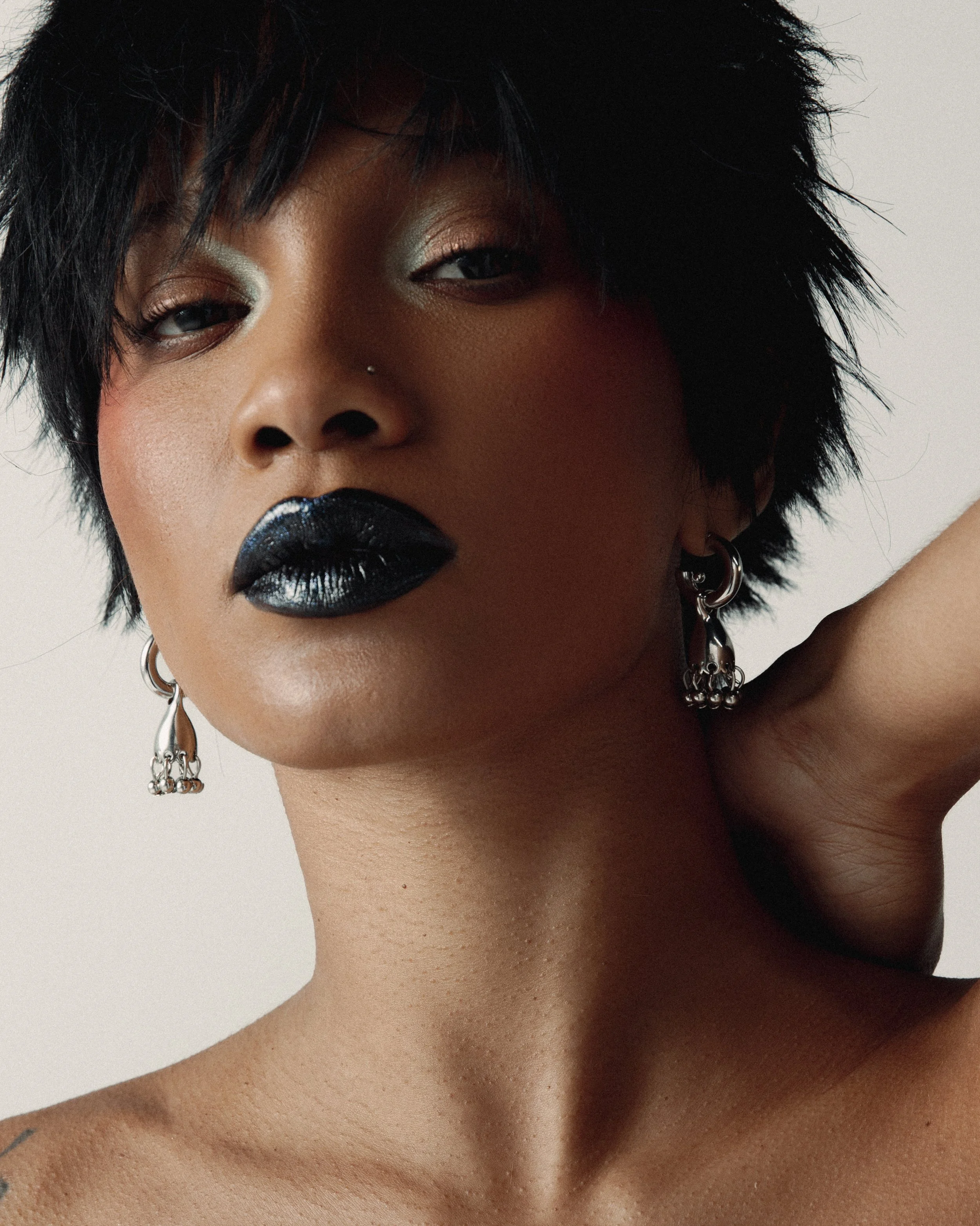 Retrato de uma mulher com pele morena, cabelo curto e volumoso, usando batom preto e maquiagem sutil, com brincos grandes de argola e piercing no nariz, contra fundo claro.