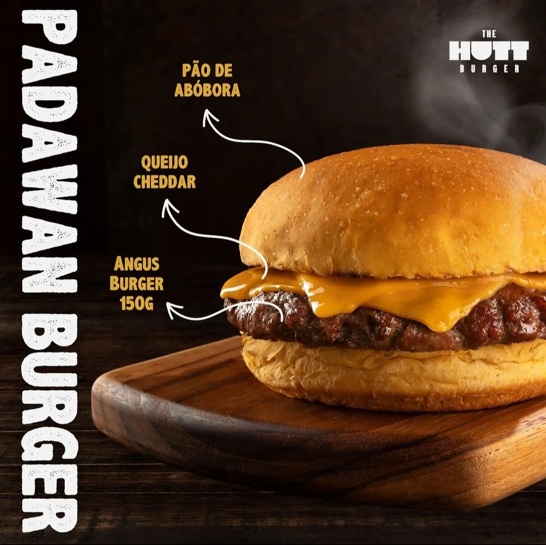 Um hambúrguer com queijo cheddar, pão de abóbora, carne de 150g de Angus, no pão servido em uma tábua de madeira escura.