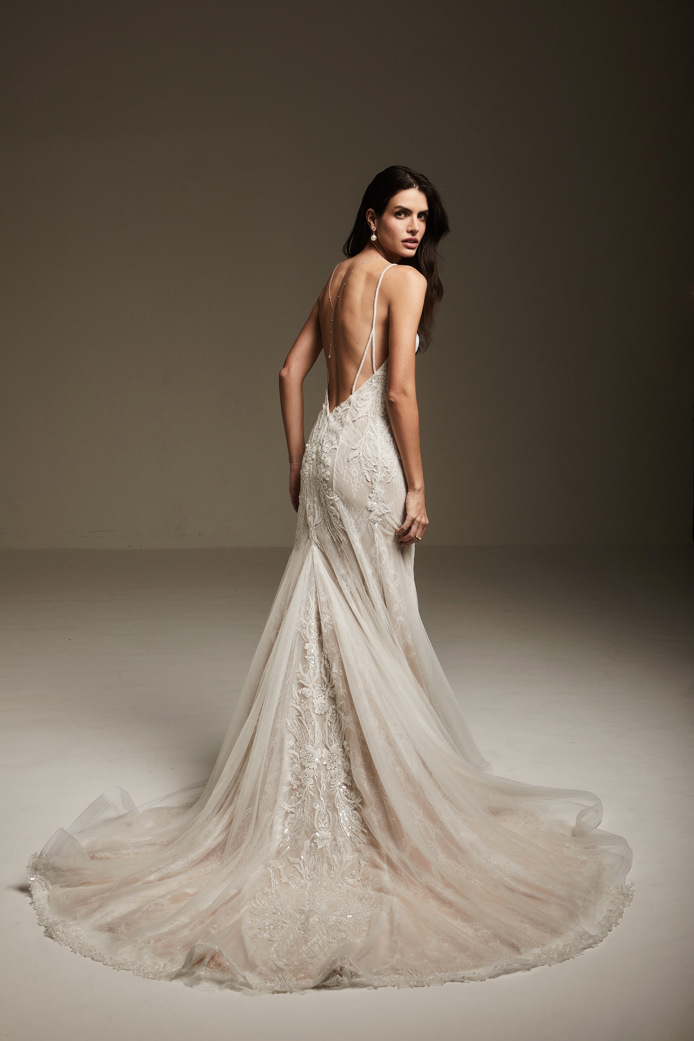 Mulher de vestido de casamento com costas expostas, cabelo escuro e ondulado, usando brincos e colar, posando em um fundo neutro.
