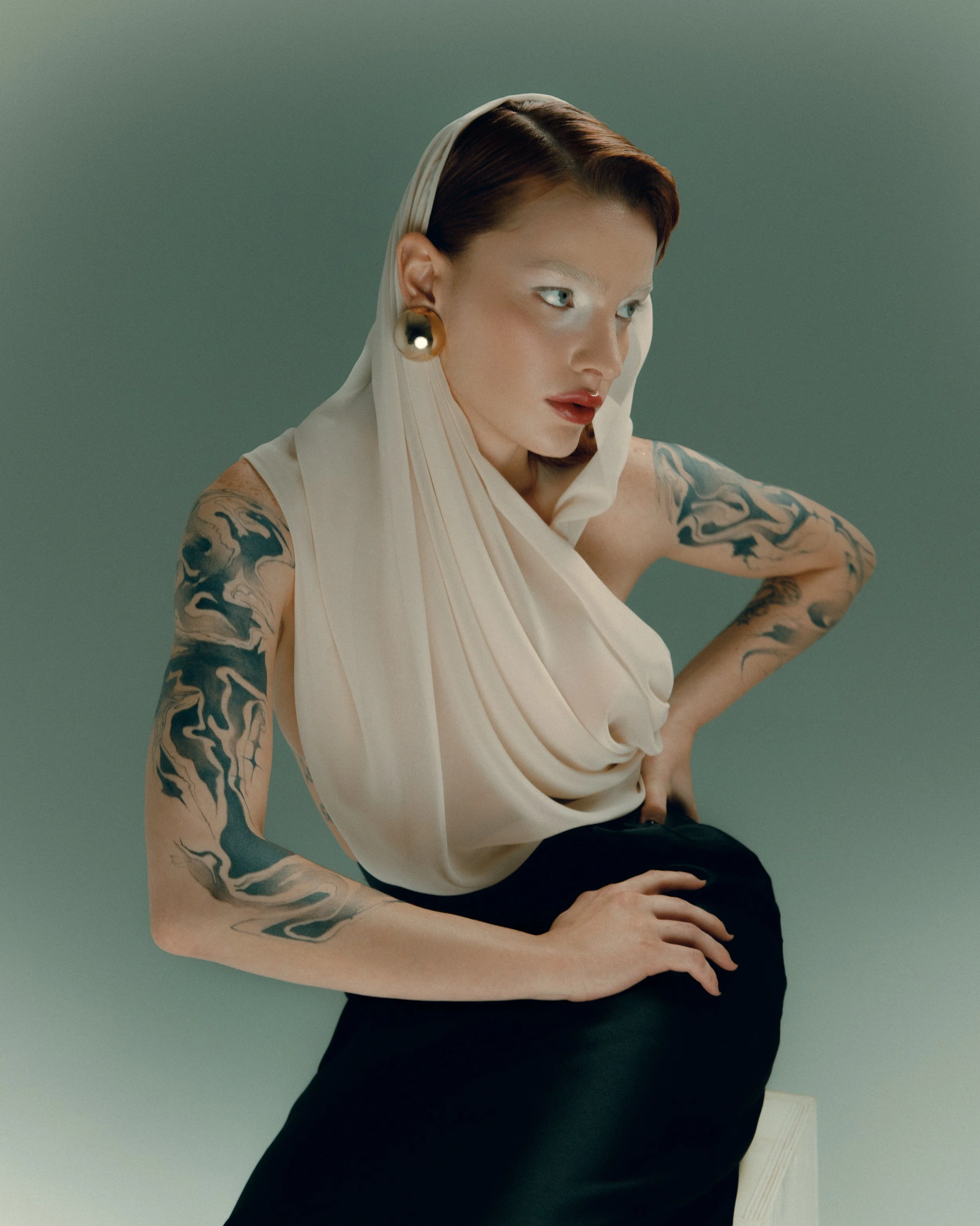 Mulher com cabelos castanho-avermelhados, vestindo blusa clara e saia preta, com tatuagens nos braços, usando brincos grandes e maquiagem com olhos claros, posando de lado contra fundo cinza.