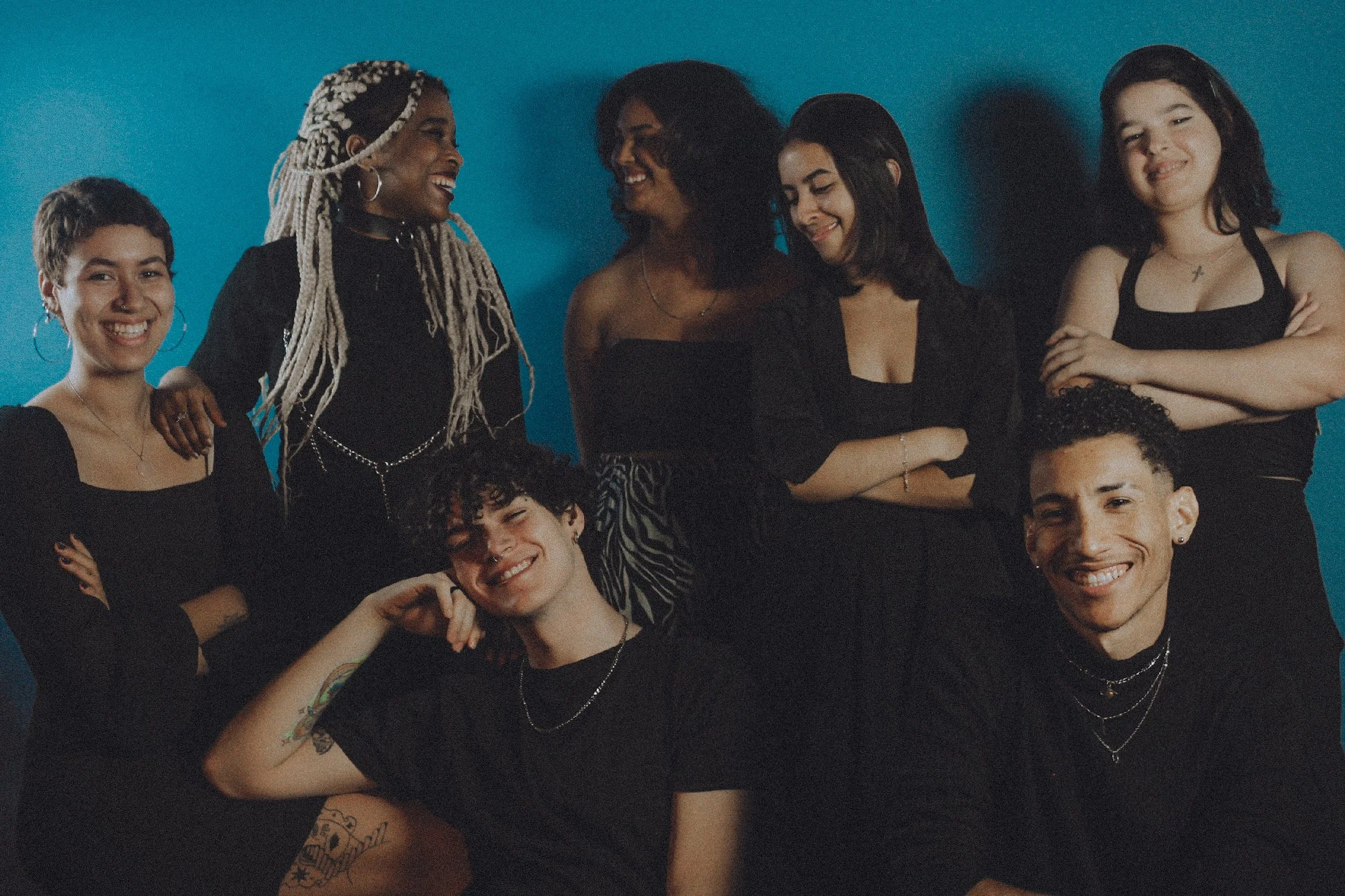 Grupo de pessoas sorrindo posando contra parede azul, usando roupas pretas, em clima de alegria e descontração.