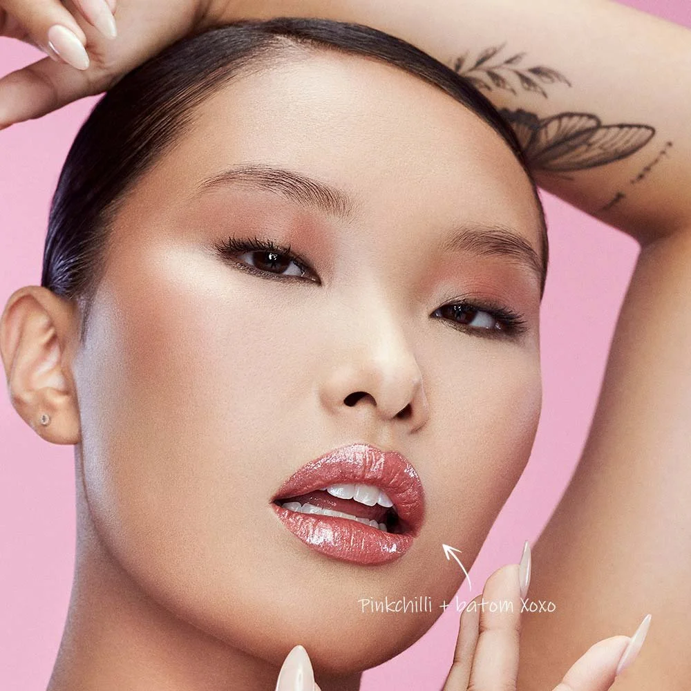 Close-up de uma mulher com maquiagem de destaque, lábios com gloss brilhante, olhando para a câmera, com fundo rosa.