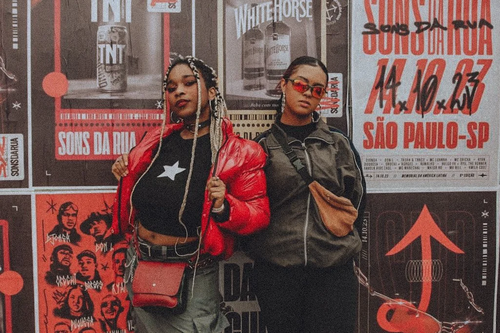 Duas jovens mulheres posando em frente a um mural com anúncios e arte urbana, uma com jaqueta vermelha e roupa preta, a outra com jaqueta escura e óculos de sol vermelhos.