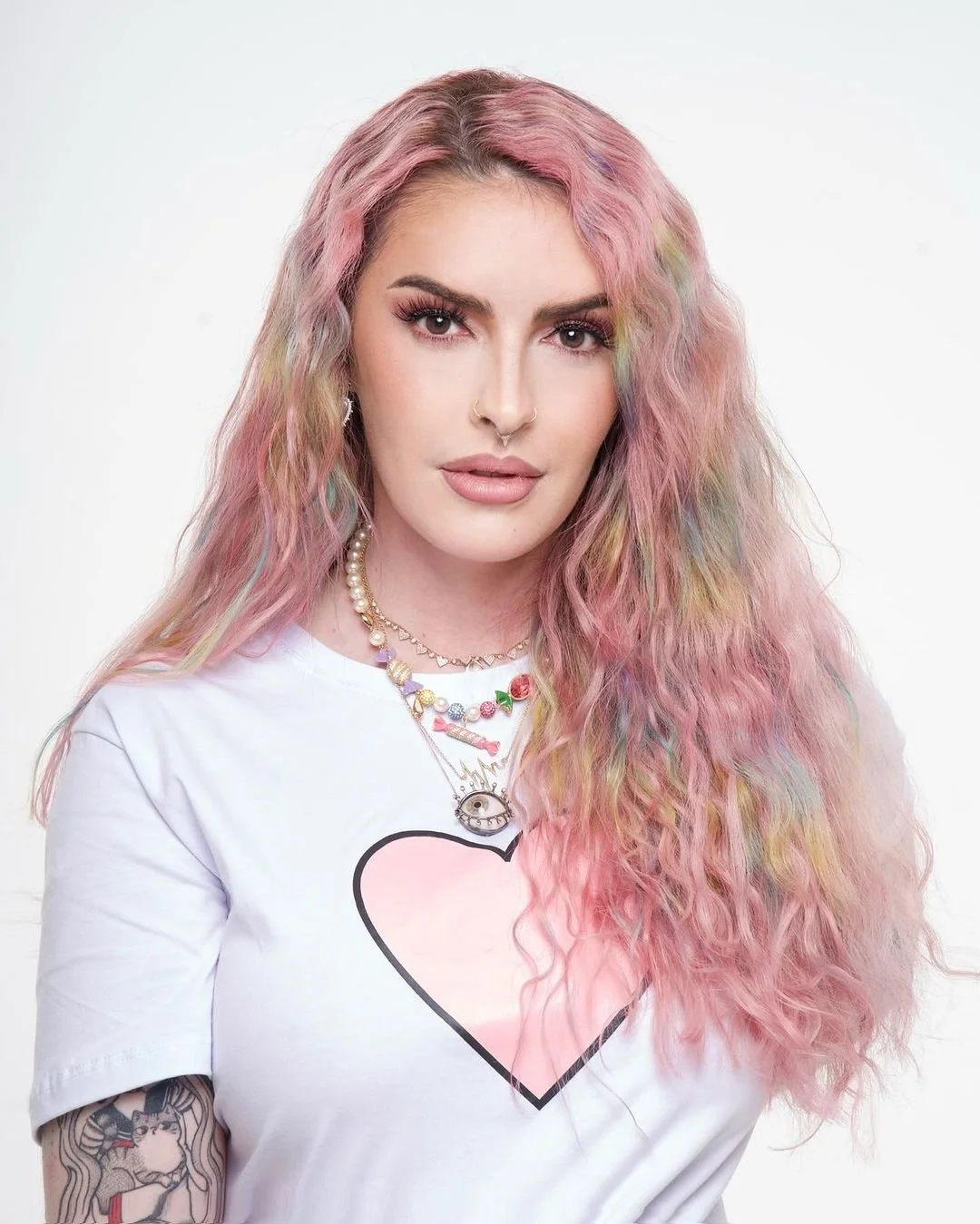 Mulher com cabelo cacheado e tingido de rosa, usando uma camiseta branca com um coração rosa na frente e diversas joias, incluindo colares e brincos, em fundo branco.
