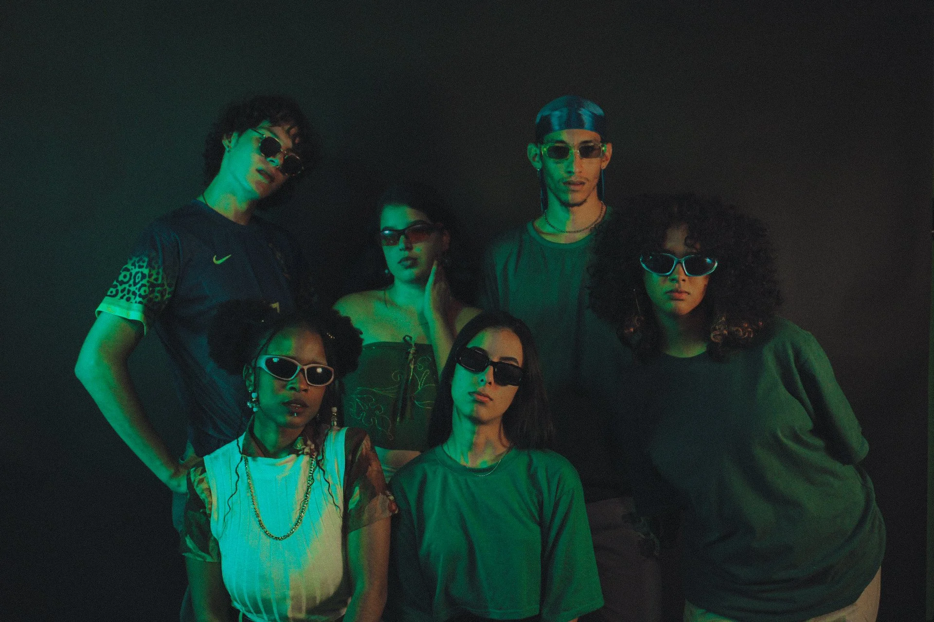 Grupo de pessoas com roupas estilosas e óculos de sol, posando numa iluminação com tons verdes e azuis escuras.