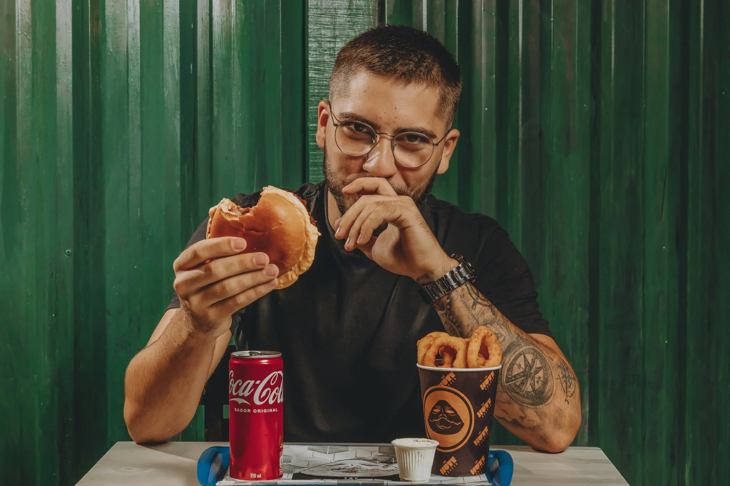 Homem com barba, óculos e tatuagens, sentado em uma mesa com uma lata de Coca-Cola, segurando um sanduíche ou pão, em um fundo de paredes verdes, com alimentos e copo de refrigerante na mesa.